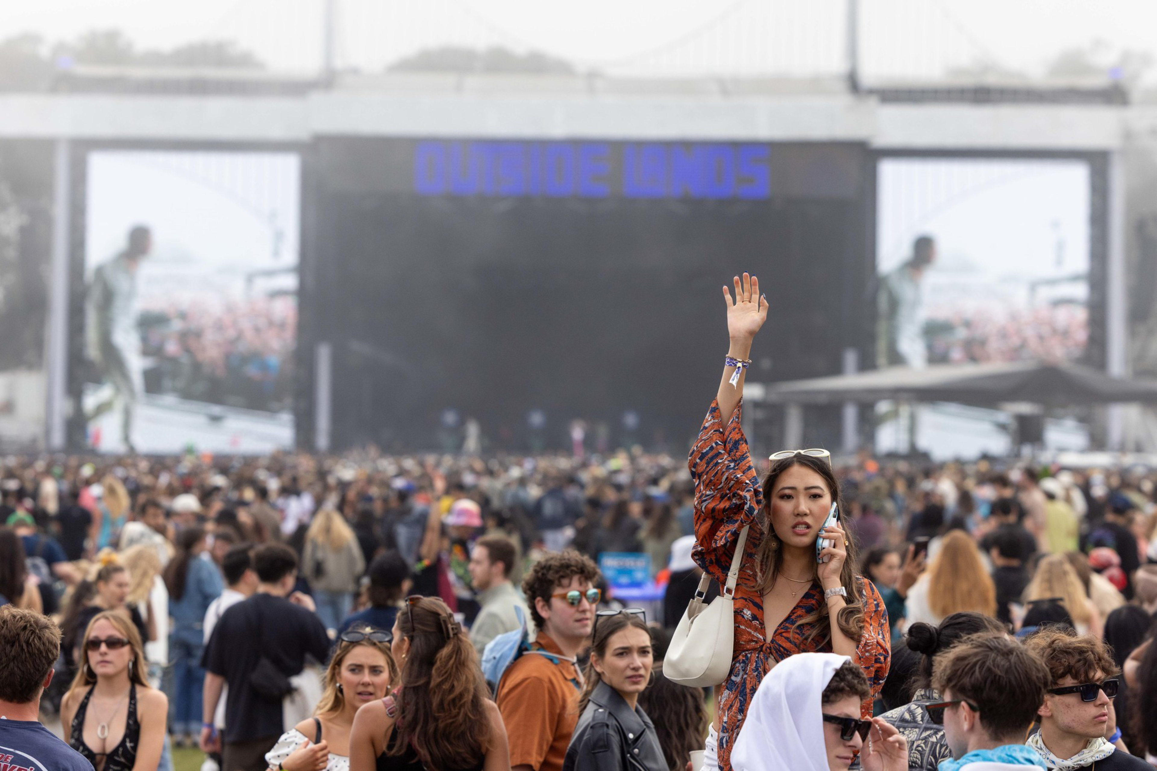 A large crowd gathers at an outdoor music festival, with a woman in patterned clothing raising her hand while talking on a phone.
