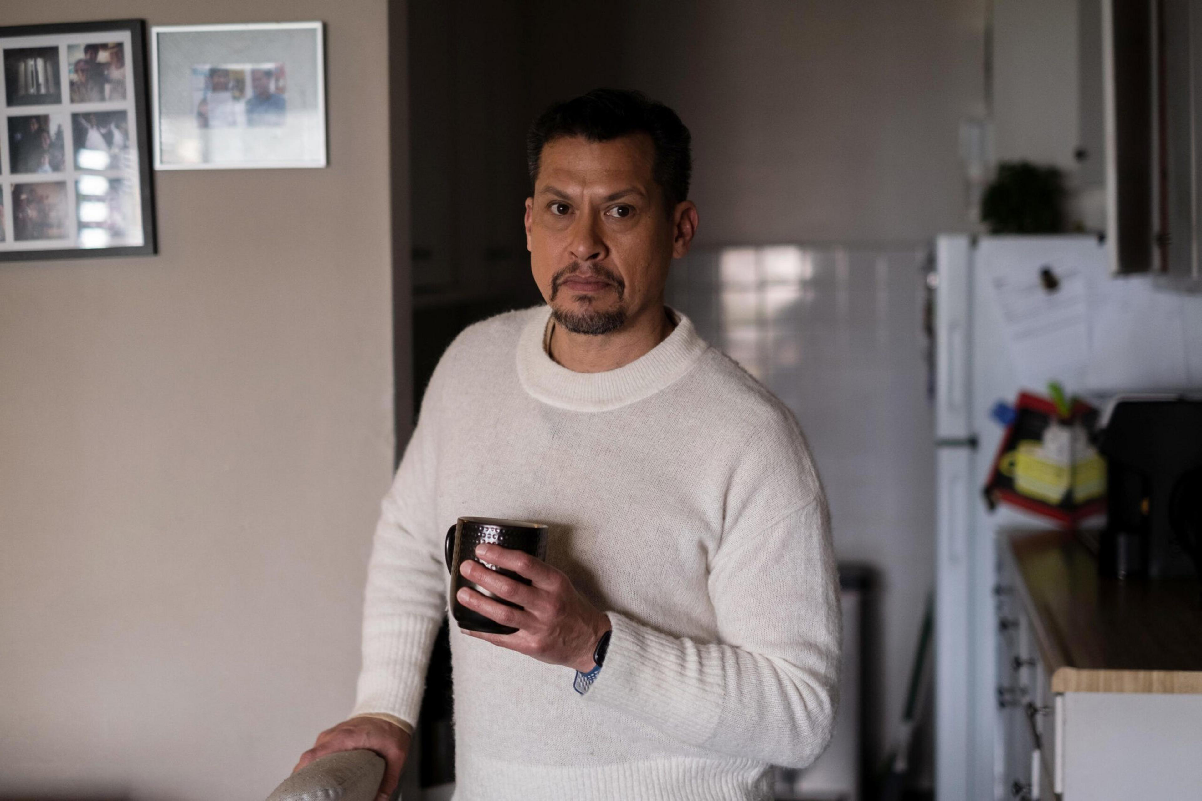 A man in a white sweater holds a black mug, standing in a kitchen with framed photos on the wall and a refrigerator in the background.