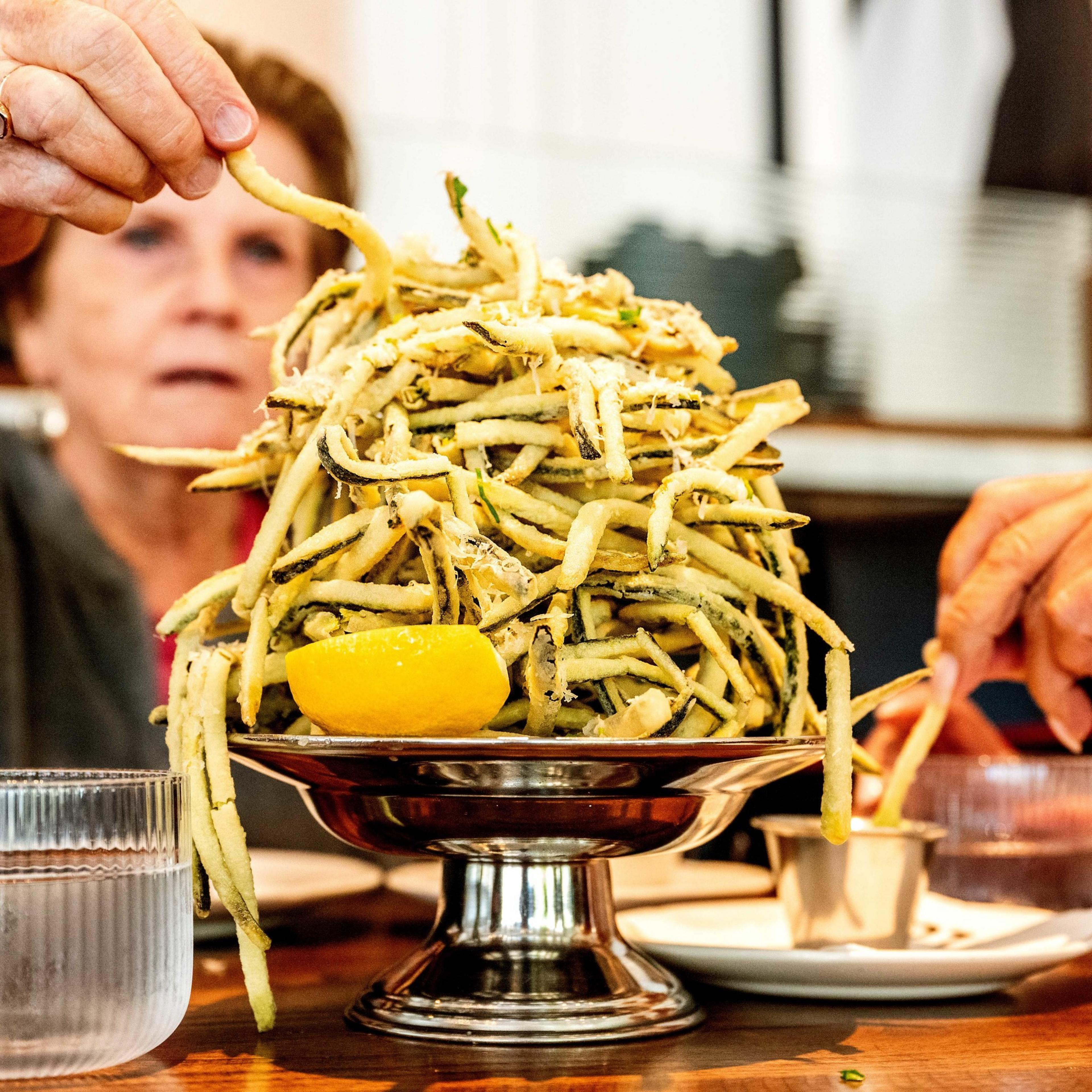 Two people share a large pile of crispy fried zucchini sticks garnished with a lemon wedge, served on a silver pedestal bowl with dipping sauces on the side.