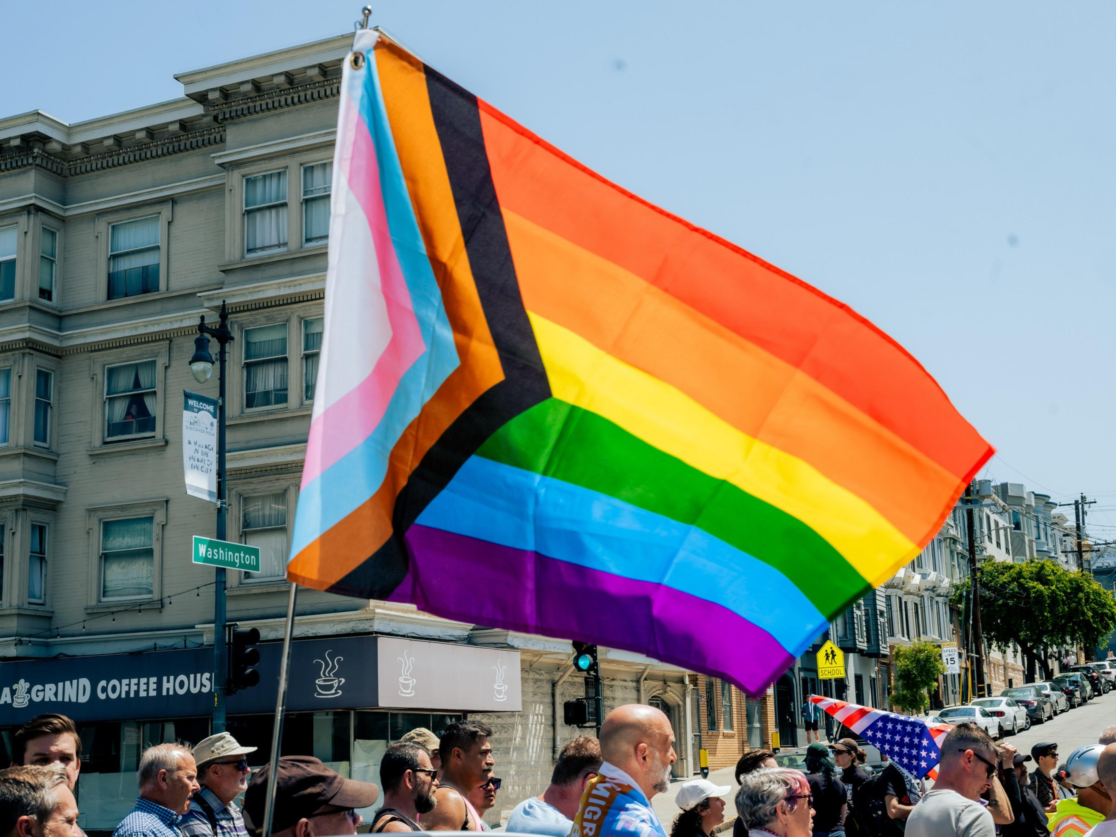 The image shows a large Progress Pride flag waving above a crowd of people standing on a city street corner, with a coffee shop and apartment building nearby.