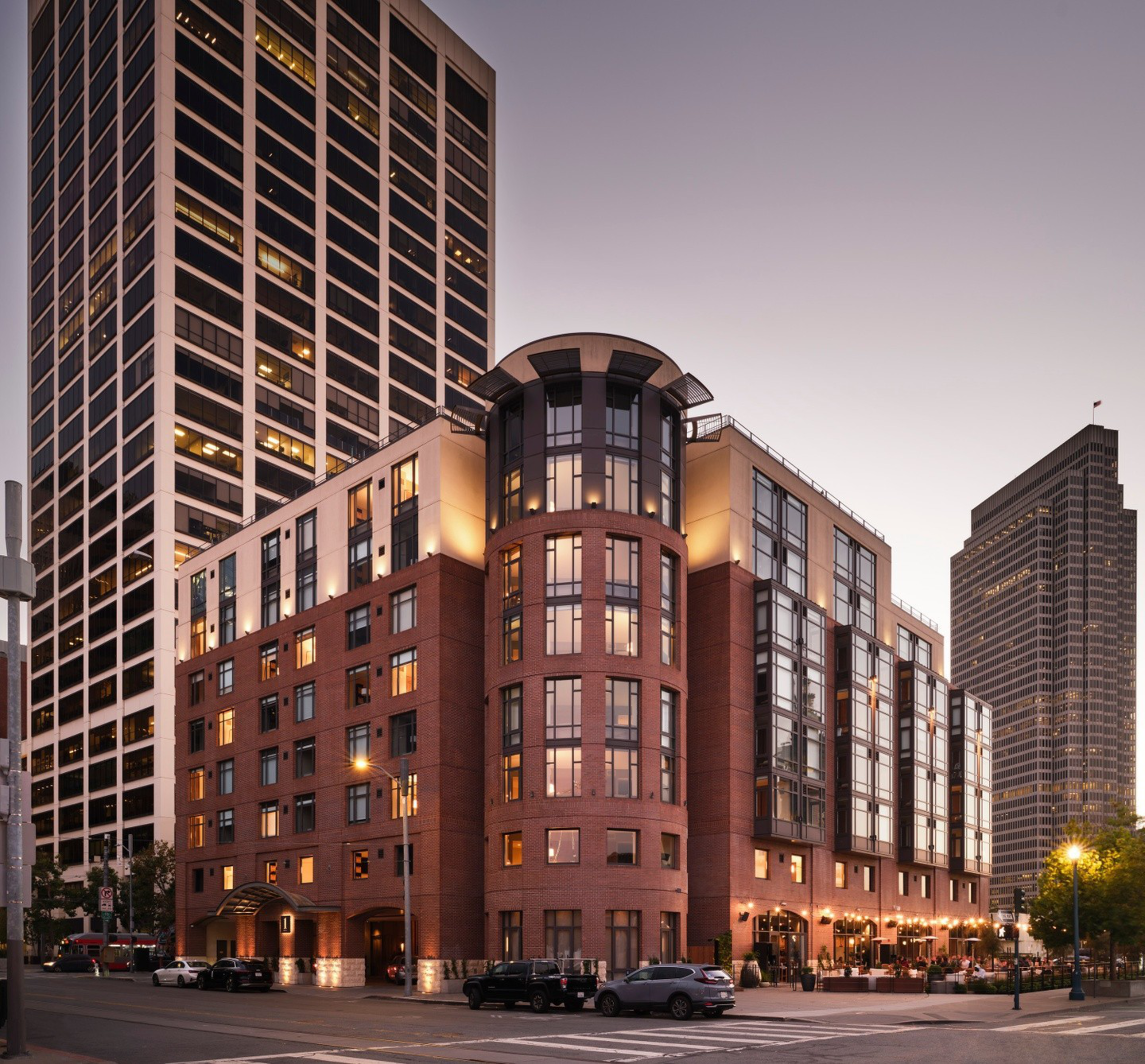 A multi-story brick and glass building with warm lights on, located at a street corner with cars parked along the road and taller buildings in the background.