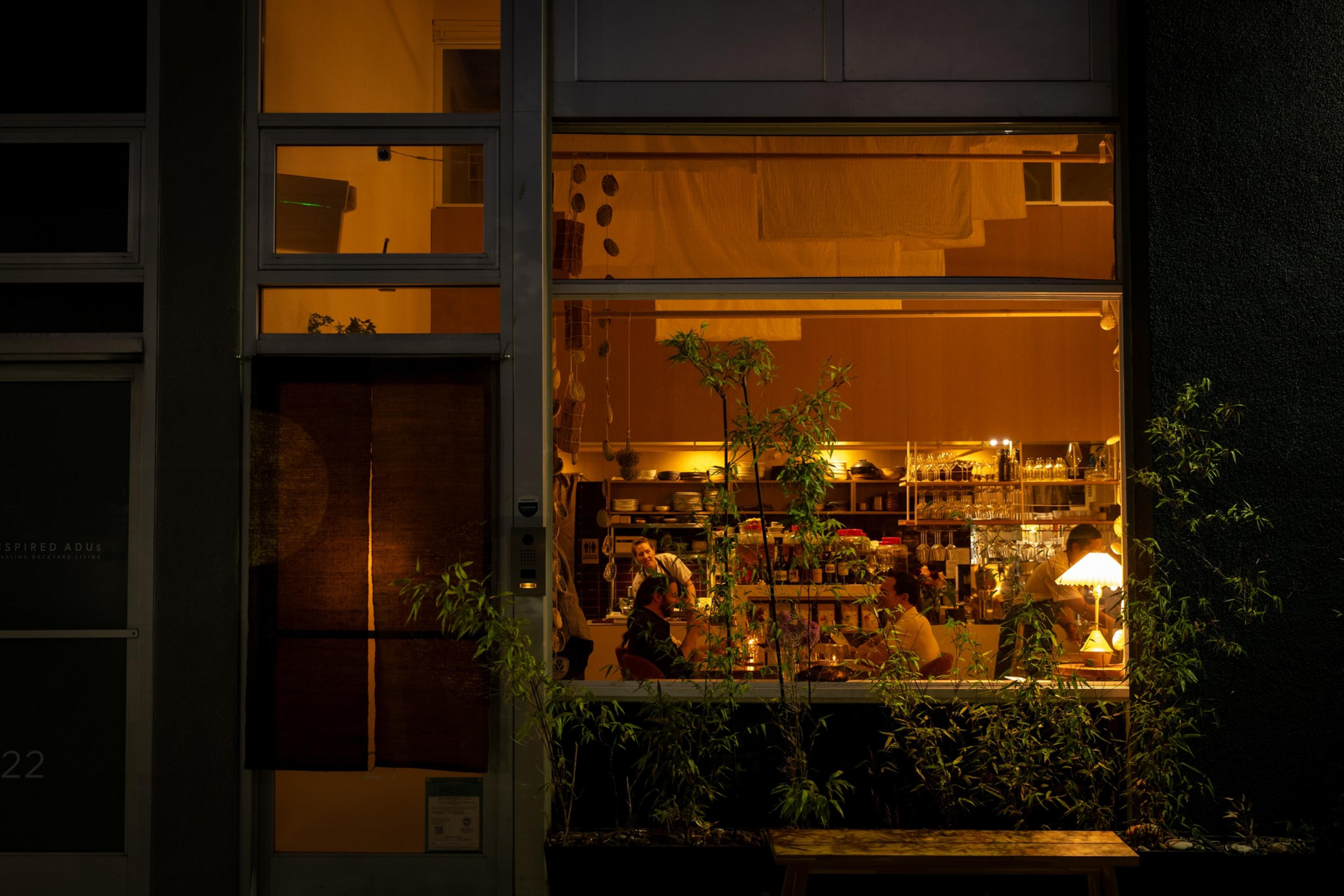 A warmly lit café with people sitting at the counter, bottles and glasses displayed on shelves, and plants outside the large window.