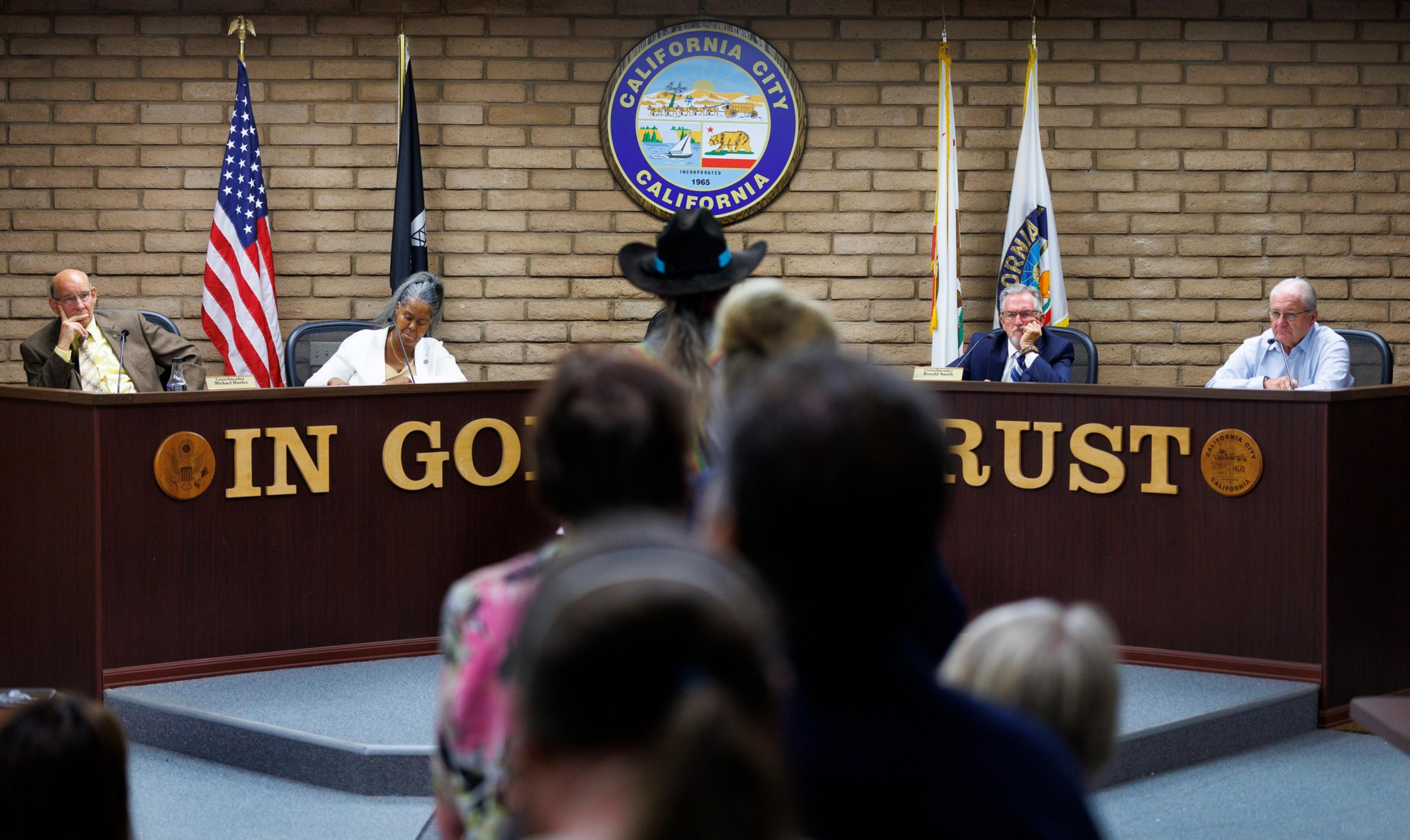 Four officials sit behind a wooden council desk with “IN GOD WE TRUST” in gold letters and a California City seal on the brick wall.