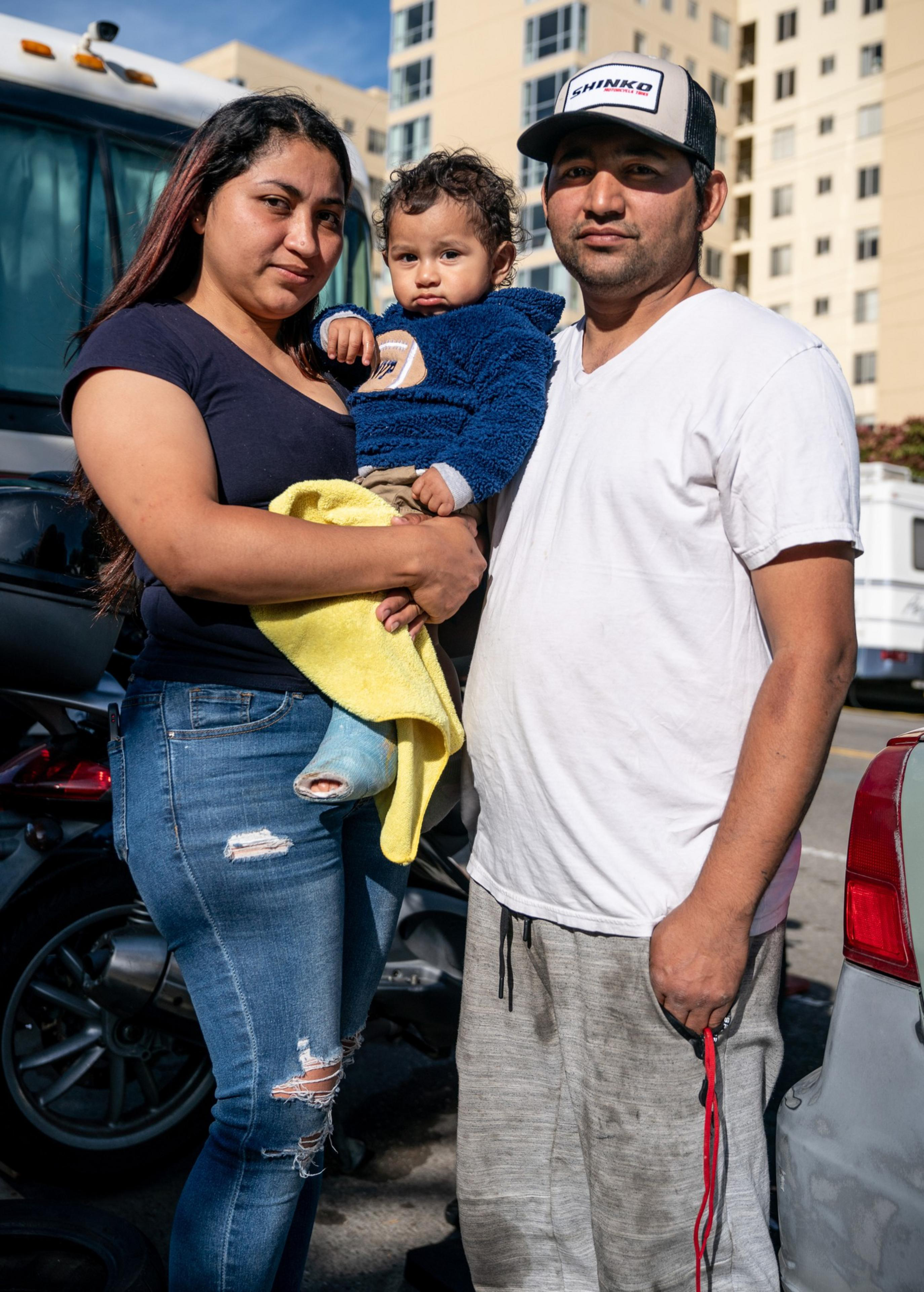 A woman holds a young child while a man stands beside them. They are outdoors in a city area with buildings, cars, and a bus in the background.