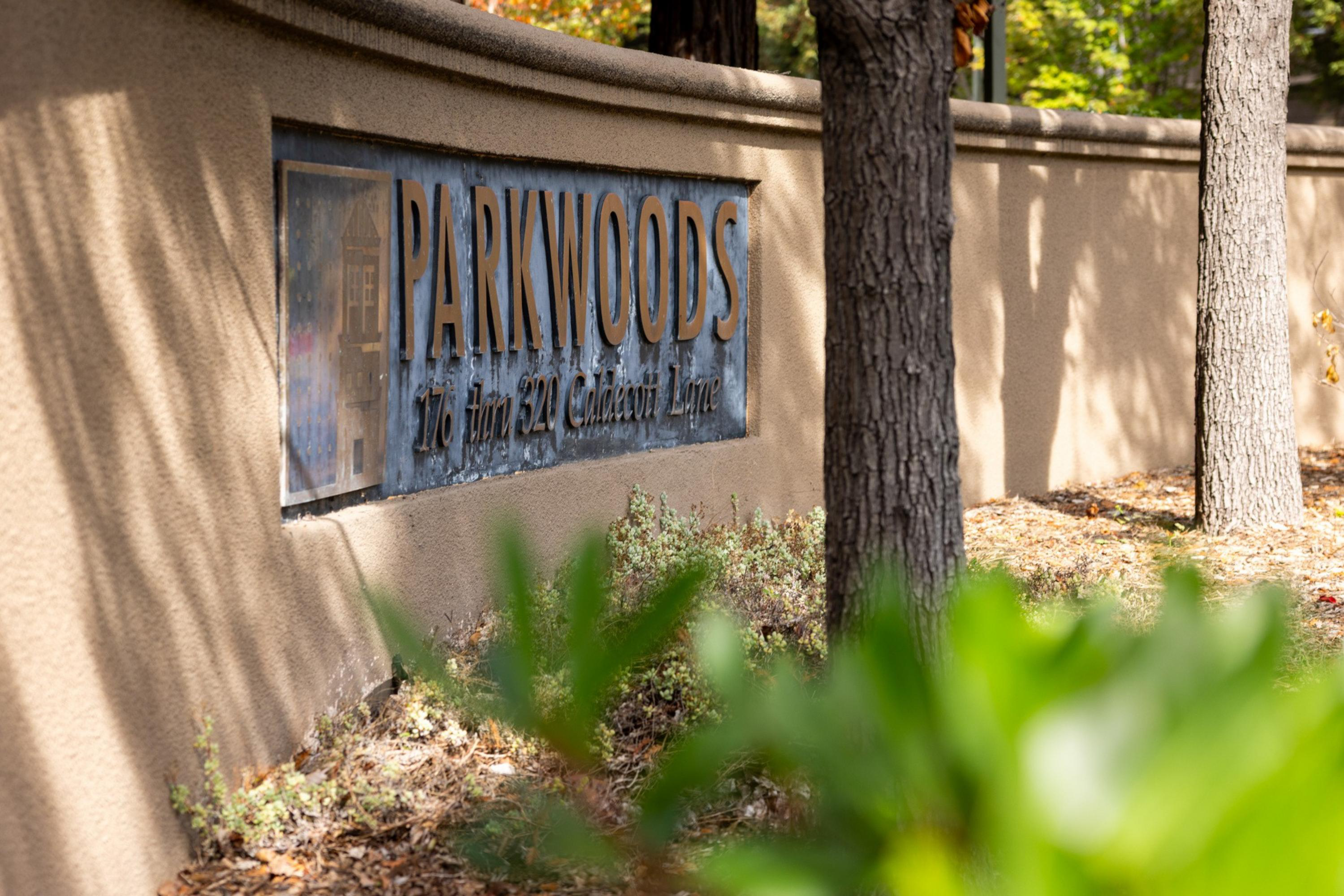 A beige stone wall with a metal sign reading “PARKWOODS 176 Jim 527 Caledon Lane” partially obscured by trees and greenery.