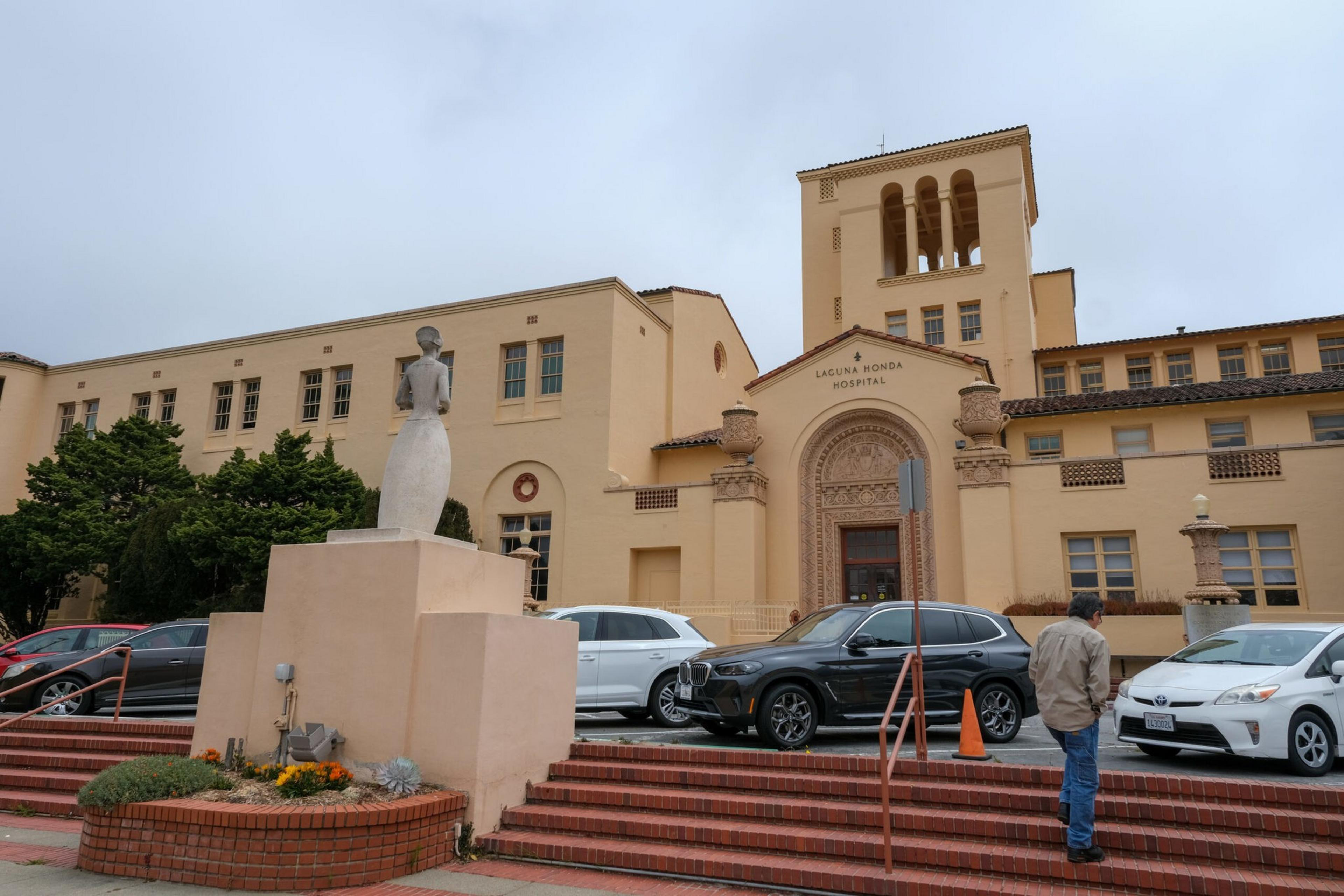 A beige hospital building with a tower and arched entrance labeled "LACUNA MONDA HOSPITAL," several parked cars, a statue on a pedestal, and a person walking nearby.