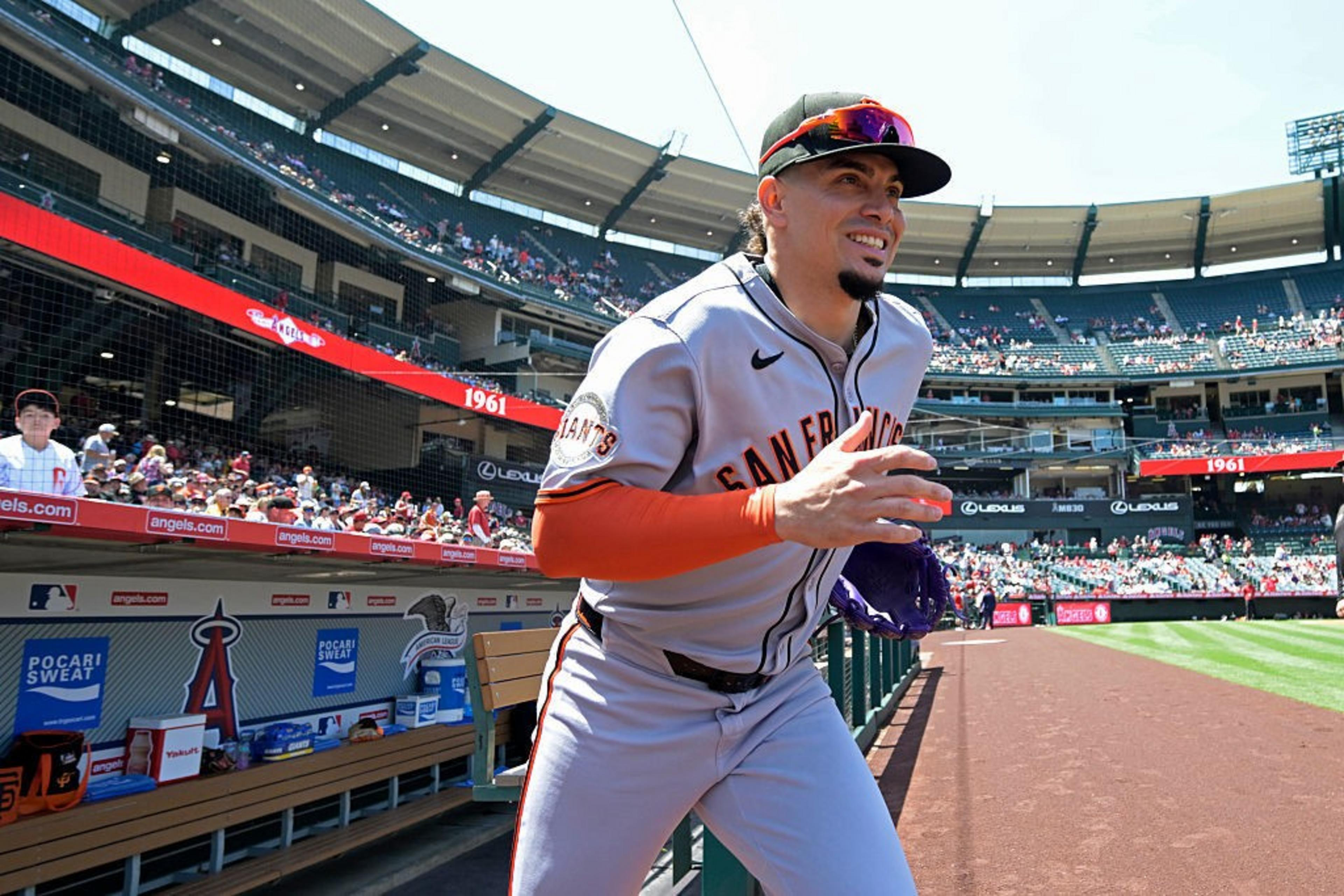 A baseball player in a San Francisco Giants uniform is running out of the dugout onto the field. The stadium is filled with fans and a bright, sunny sky is overhead.