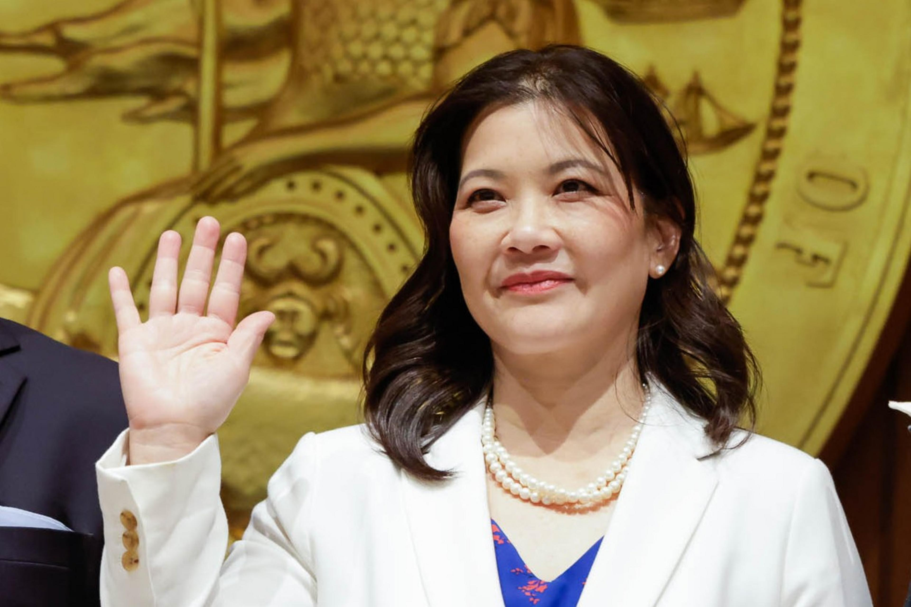 A woman in a white outfit smiling and waving, with ornate decor in the background.
