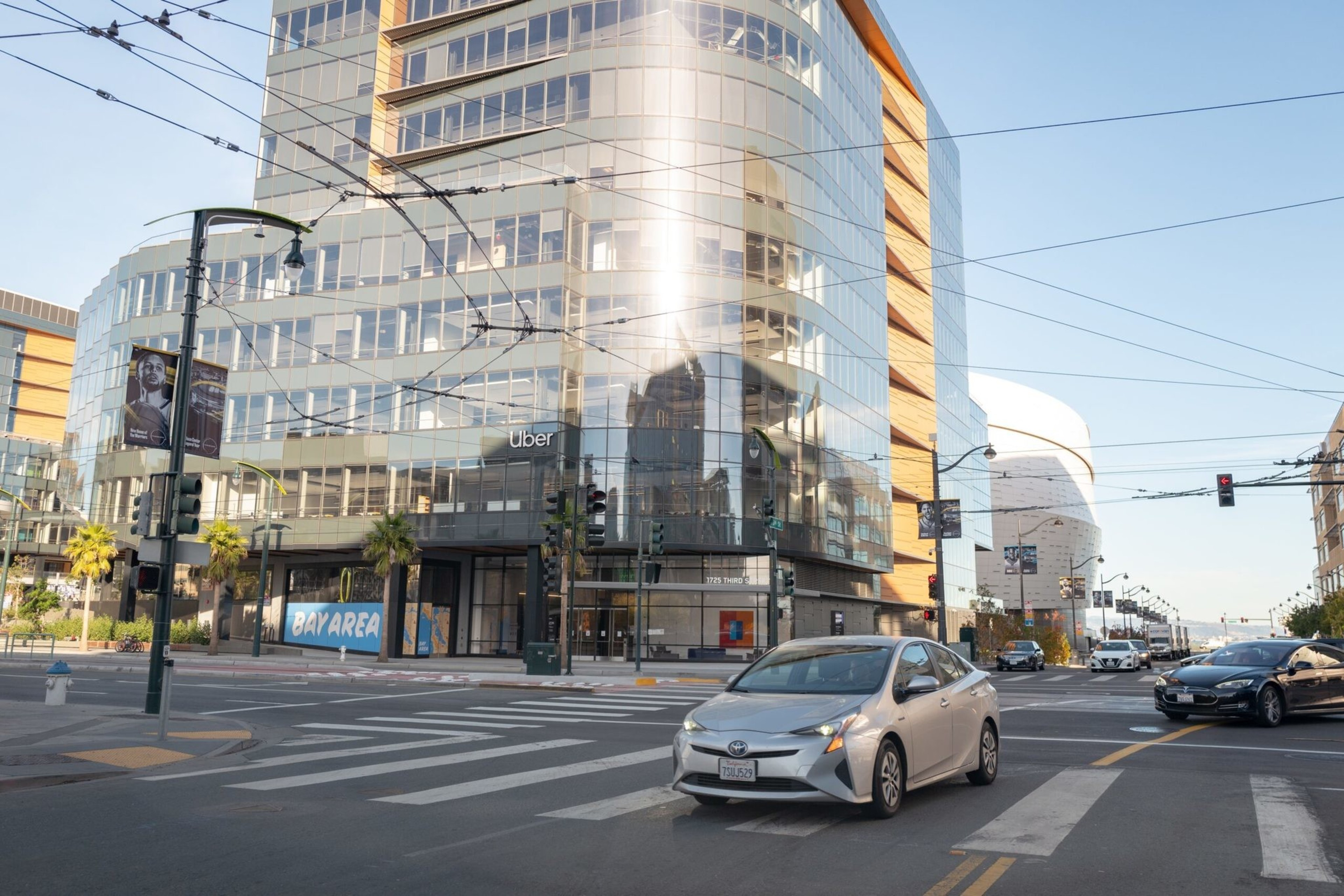 A modern glass office building with "Uber" signage sits at an urban intersection with a silver car driving through and clear blue skies above.