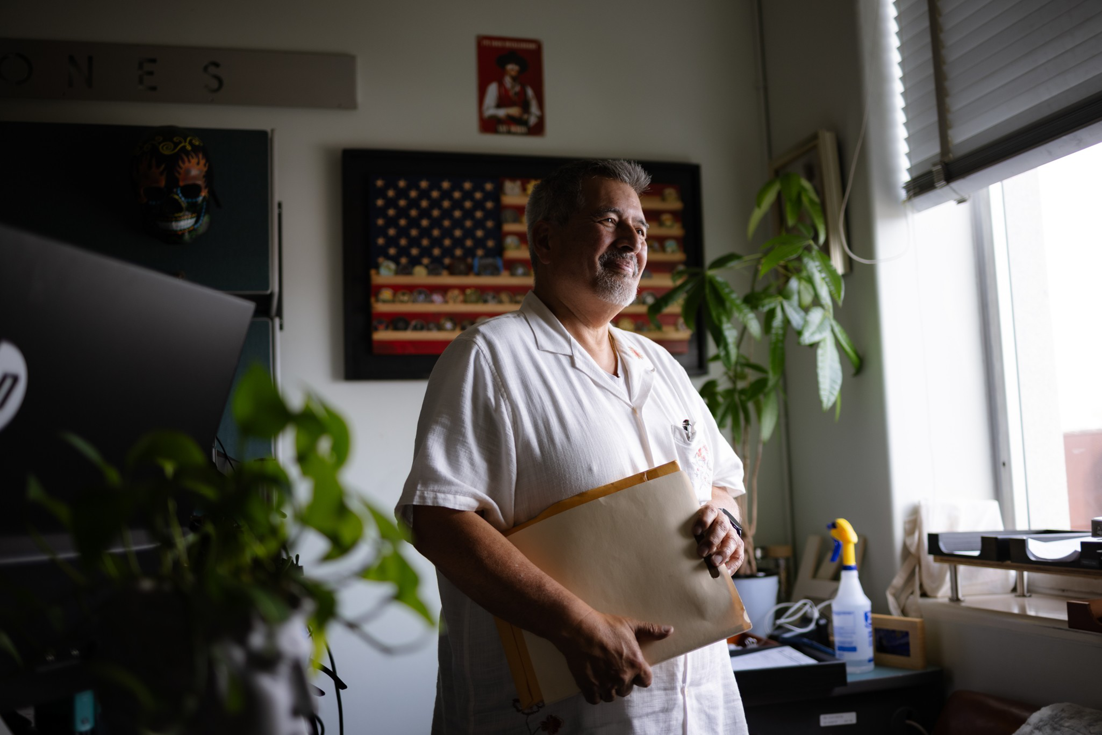 A smiling man in a white shirt holds a large envelope, standing near a window with plants and an American flag painting behind him.