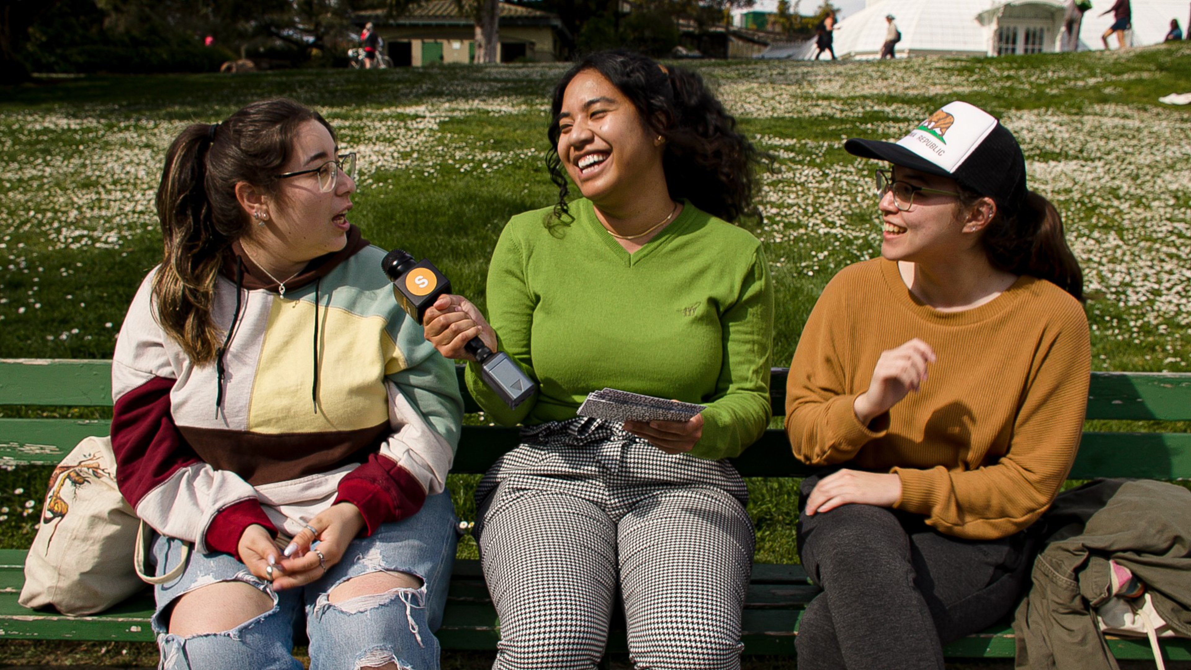 Three young women sit on a green bench outdoors; one in the middle holds a microphone and notebook, interviewing the others who are smiling and talking.