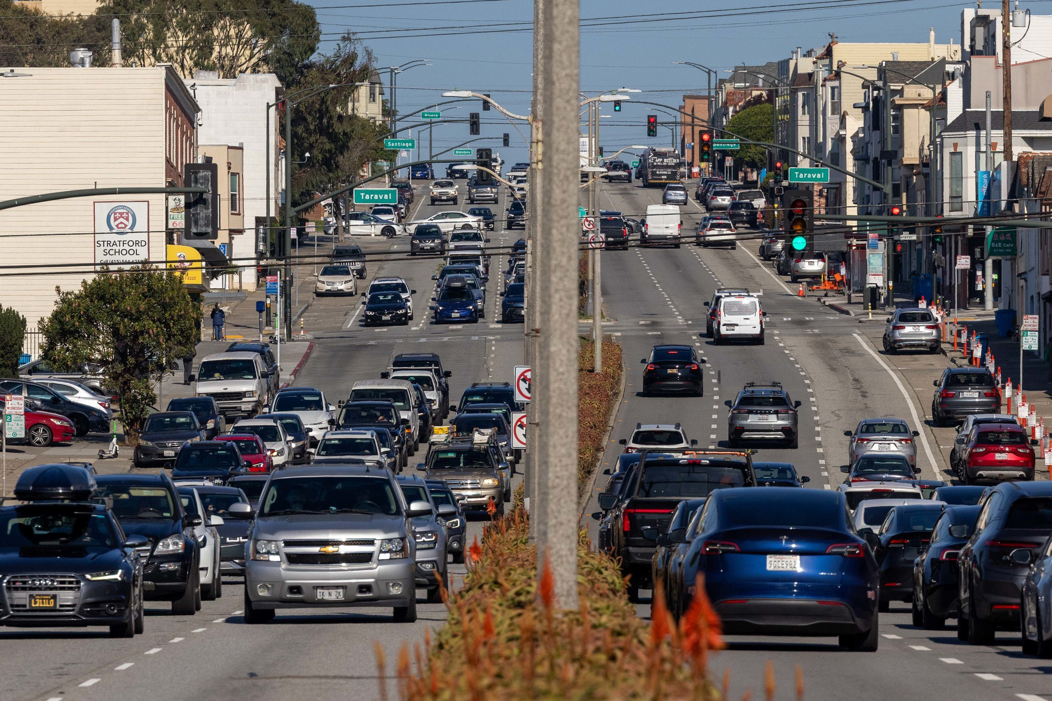 A busy city street with multiple lanes of cars, some stopped at a red light and others moving forward, with buildings and street signs on both sides.