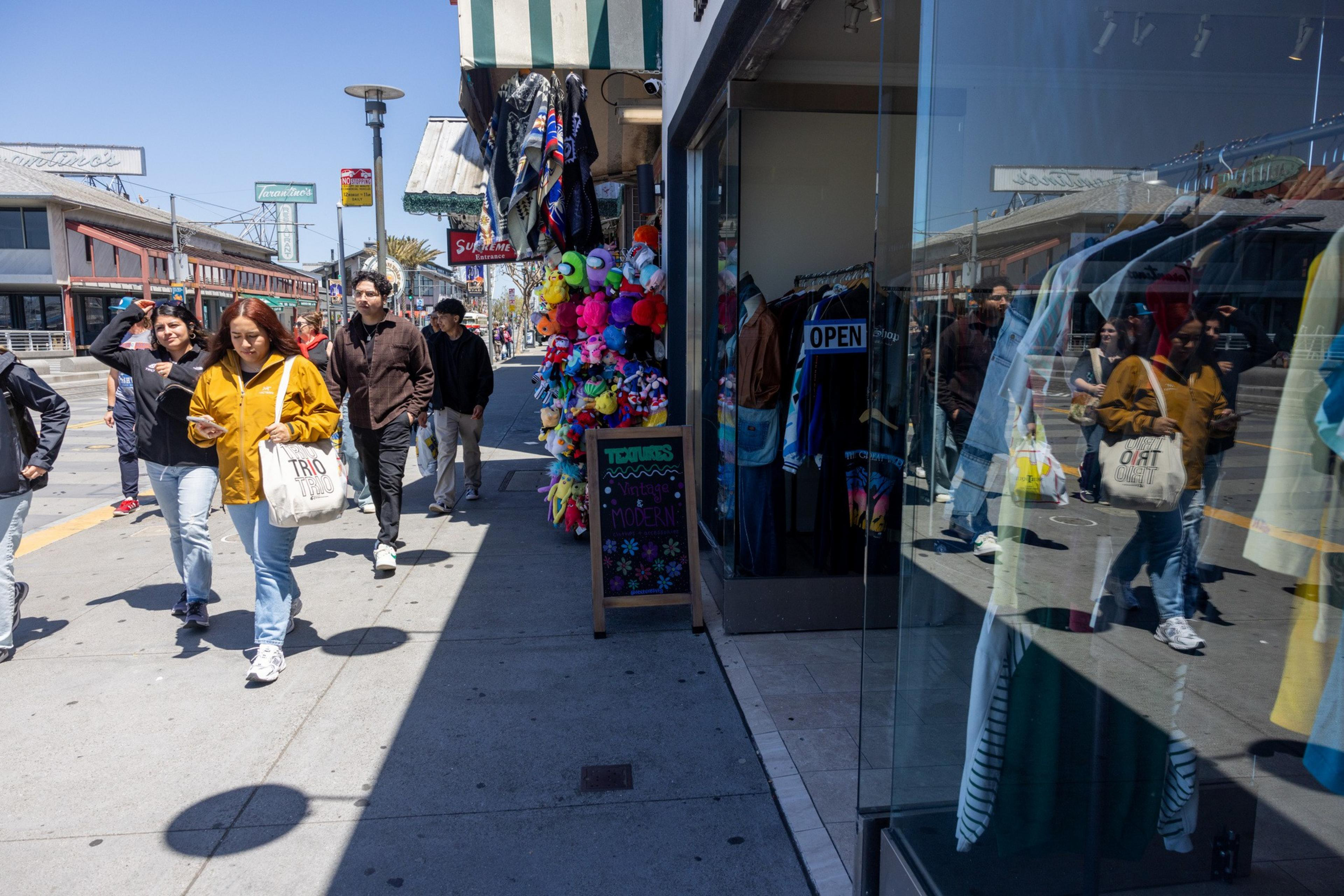 A busy street scene shows people walking past shops, including one with a colorful display of stuffed animals and a sign reading &quot;TOPS Vintage Modern,&quot; and another with an &quot;OPEN&quot; sign.