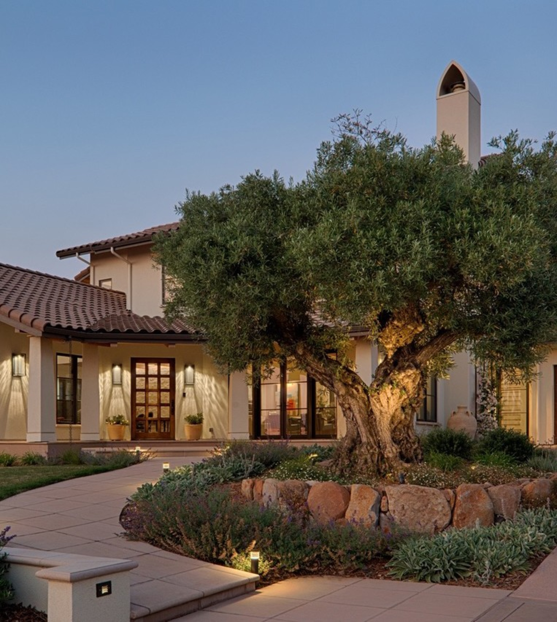 A Mediterranean-style house with a tiled roof is seen at dusk. The entrance has lit pathways and an old olive tree surrounded by low shrubs and rocks.