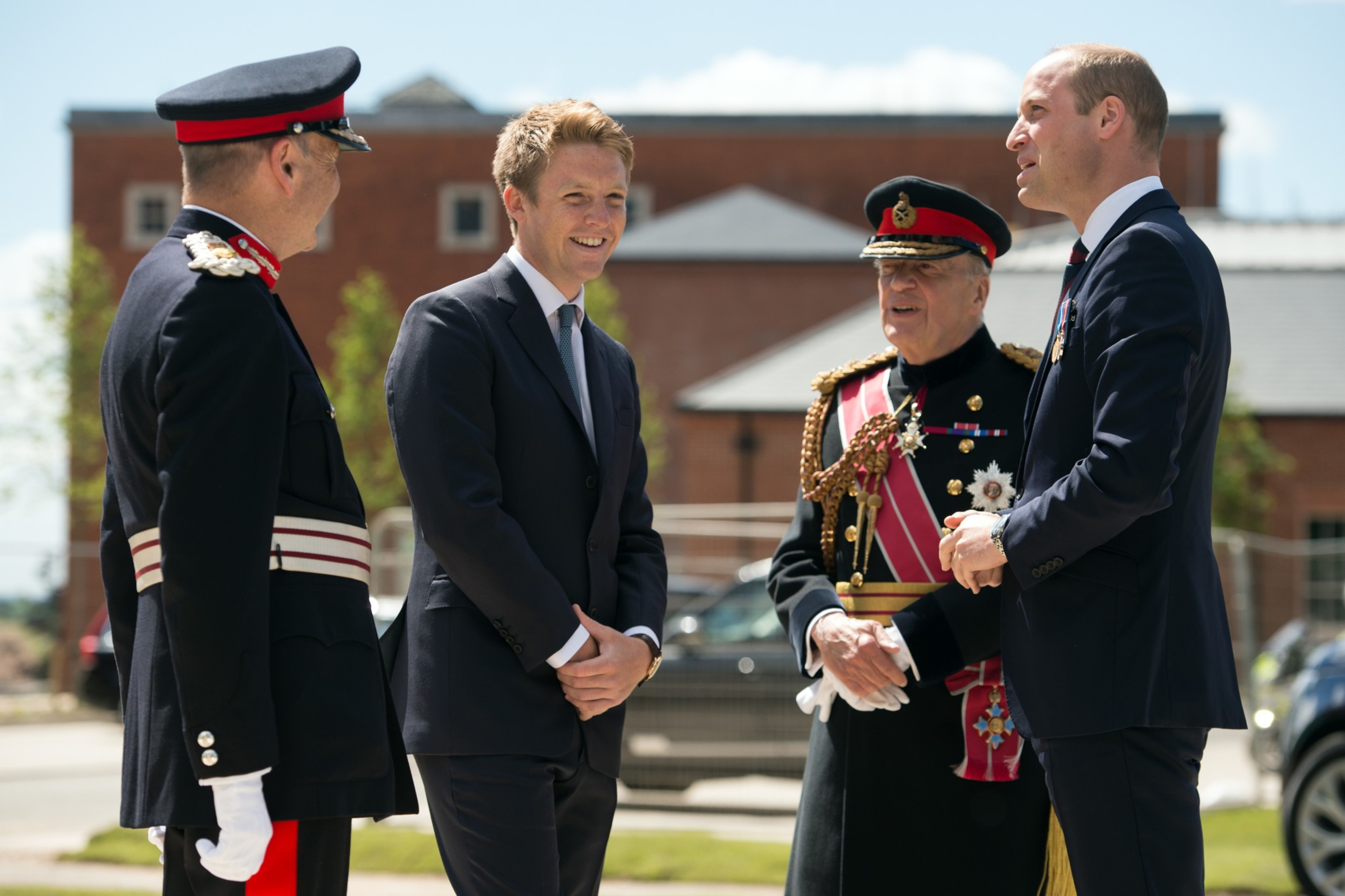 Two men in formal suits and two men in decorated military uniforms are engaged in conversation outdoors near a building on a sunny day.