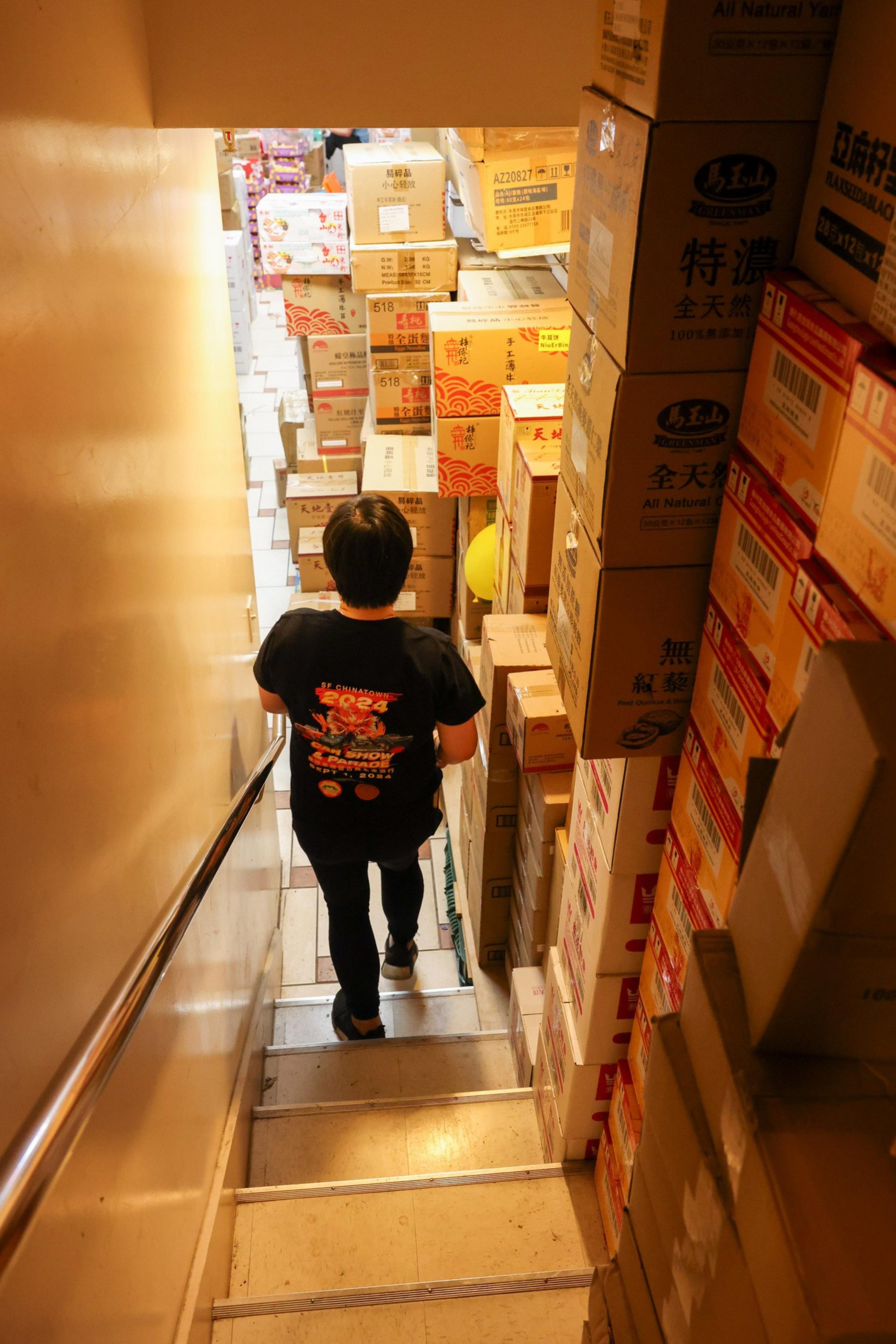 A person walks down a narrow staircase lined with stacks of cardboard boxes on both sides, leading to a cluttered basement.