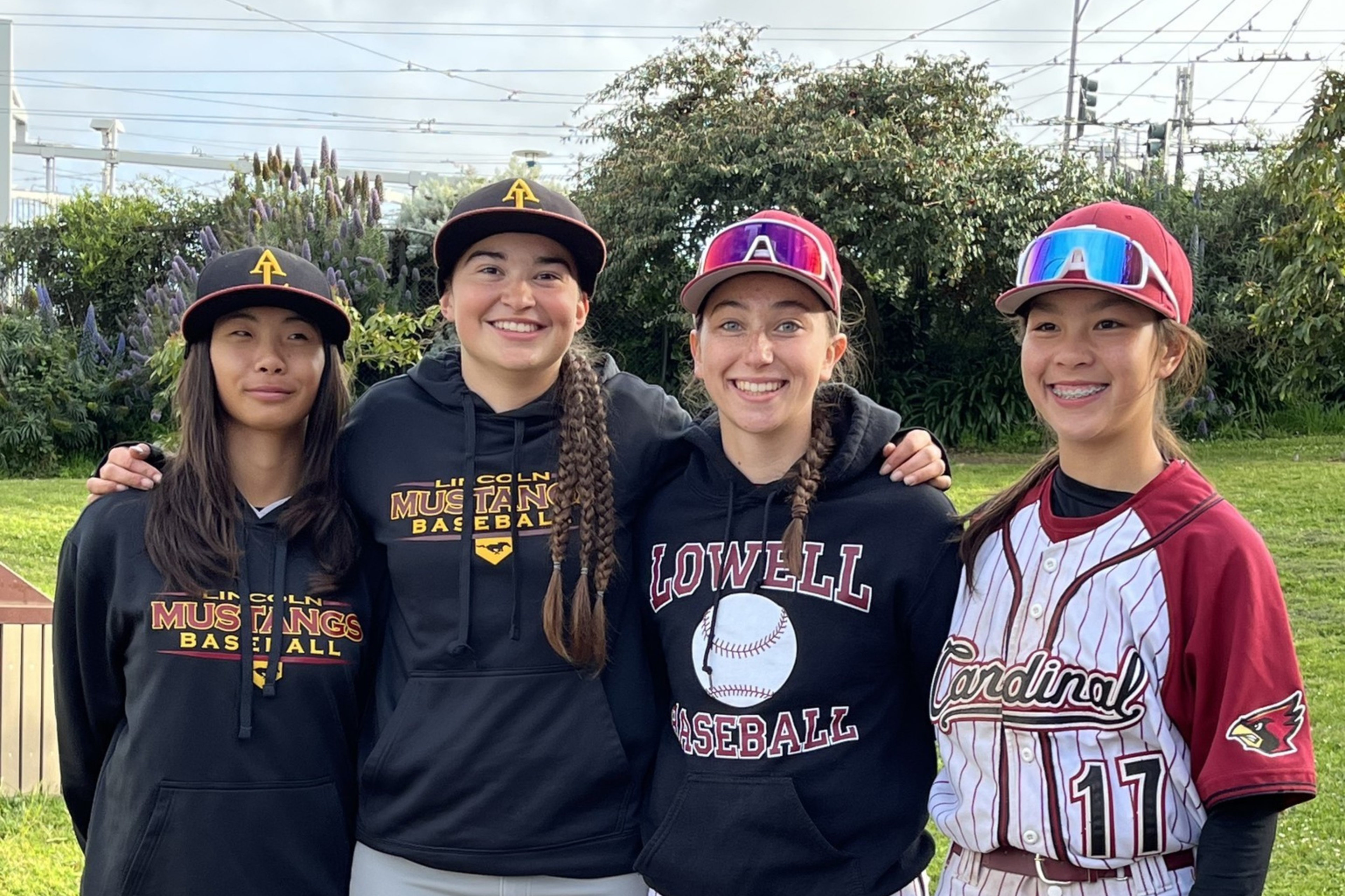 Four young female baseball players stand together outside, wearing uniforms and hoodies from different teams, smiling with arms around each other.
