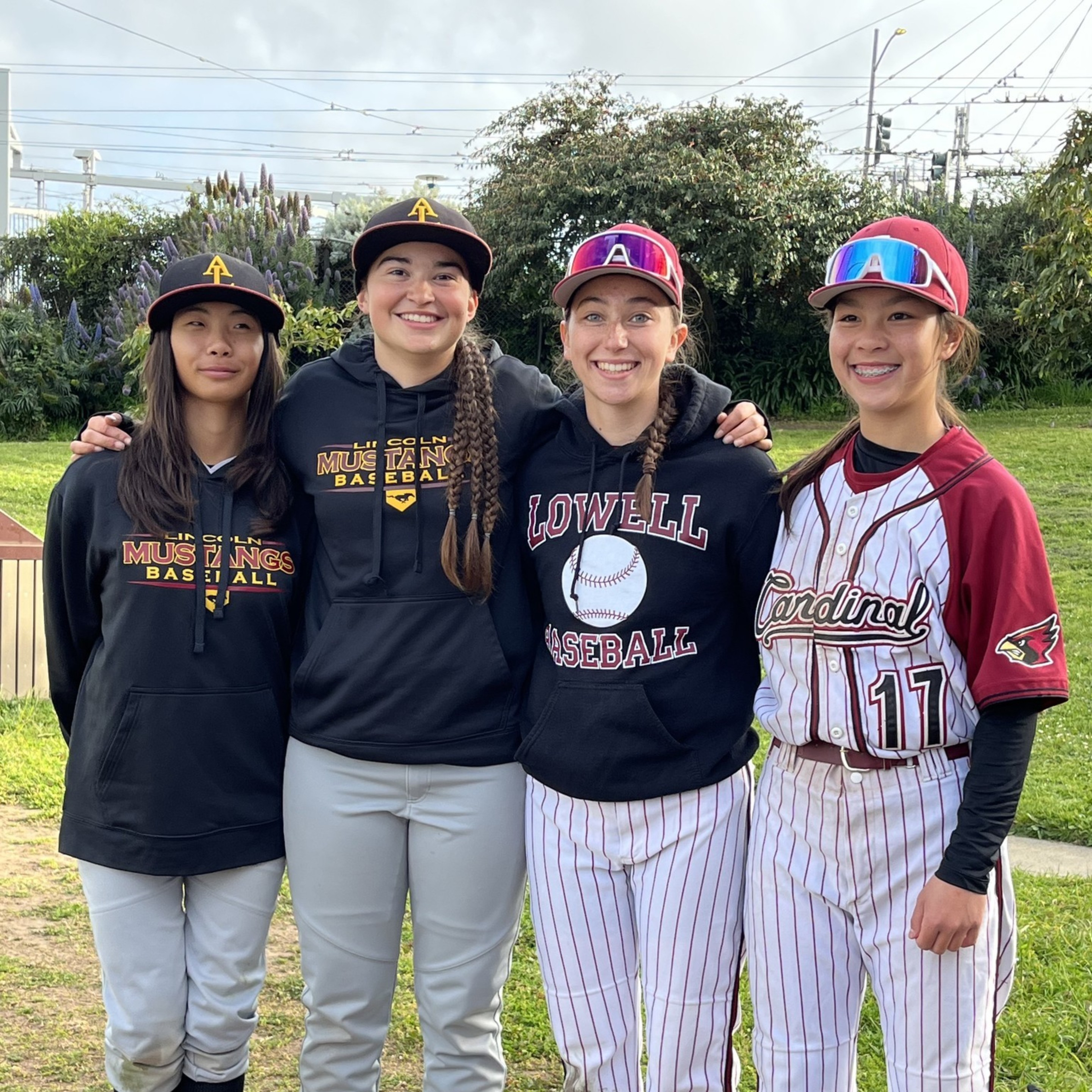 Four young female baseball players stand together outside, wearing uniforms and hoodies from different teams, smiling with arms around each other.