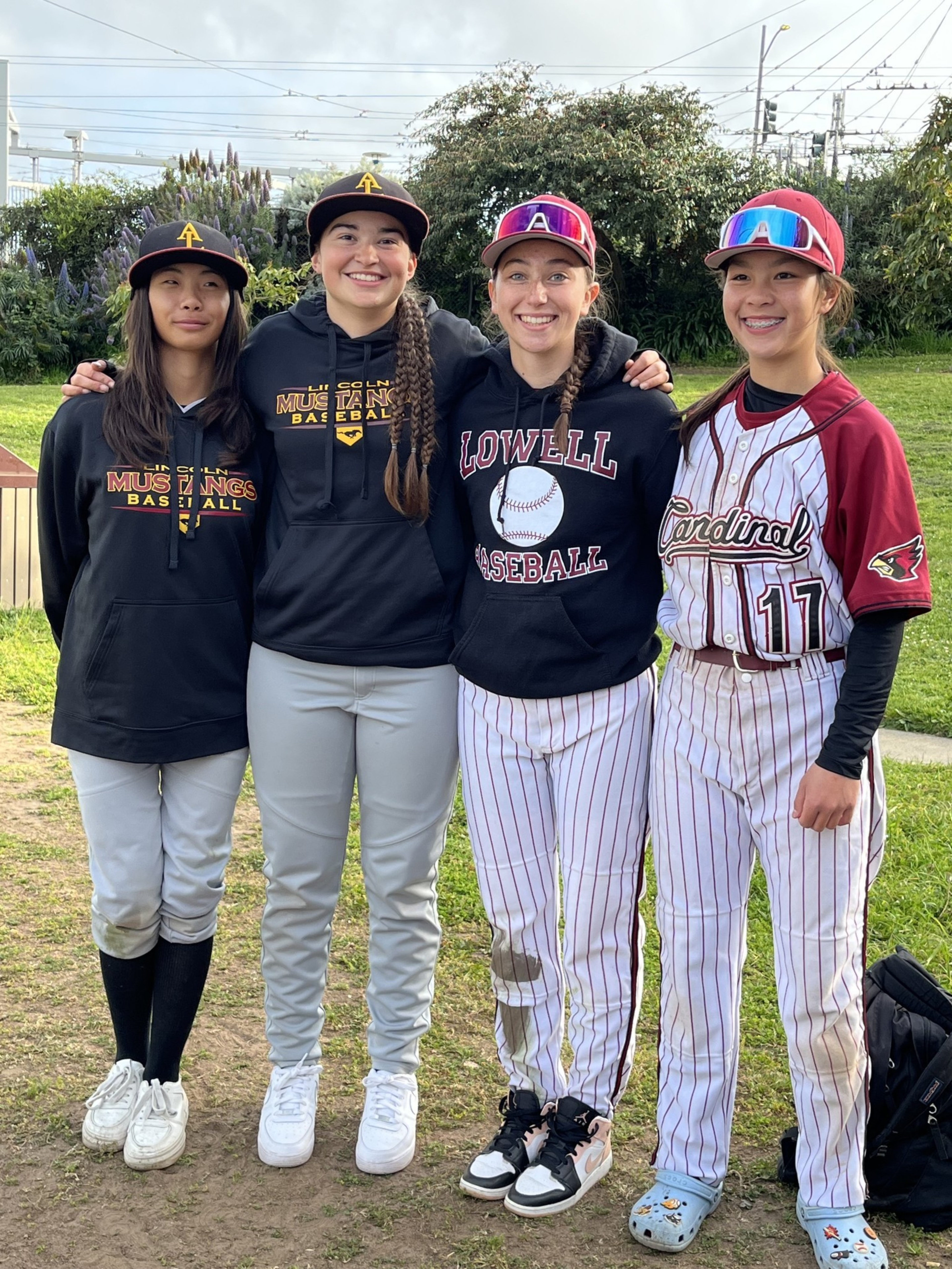 Four young female baseball players stand together outside, wearing uniforms and hoodies from different teams, smiling with arms around each other.
