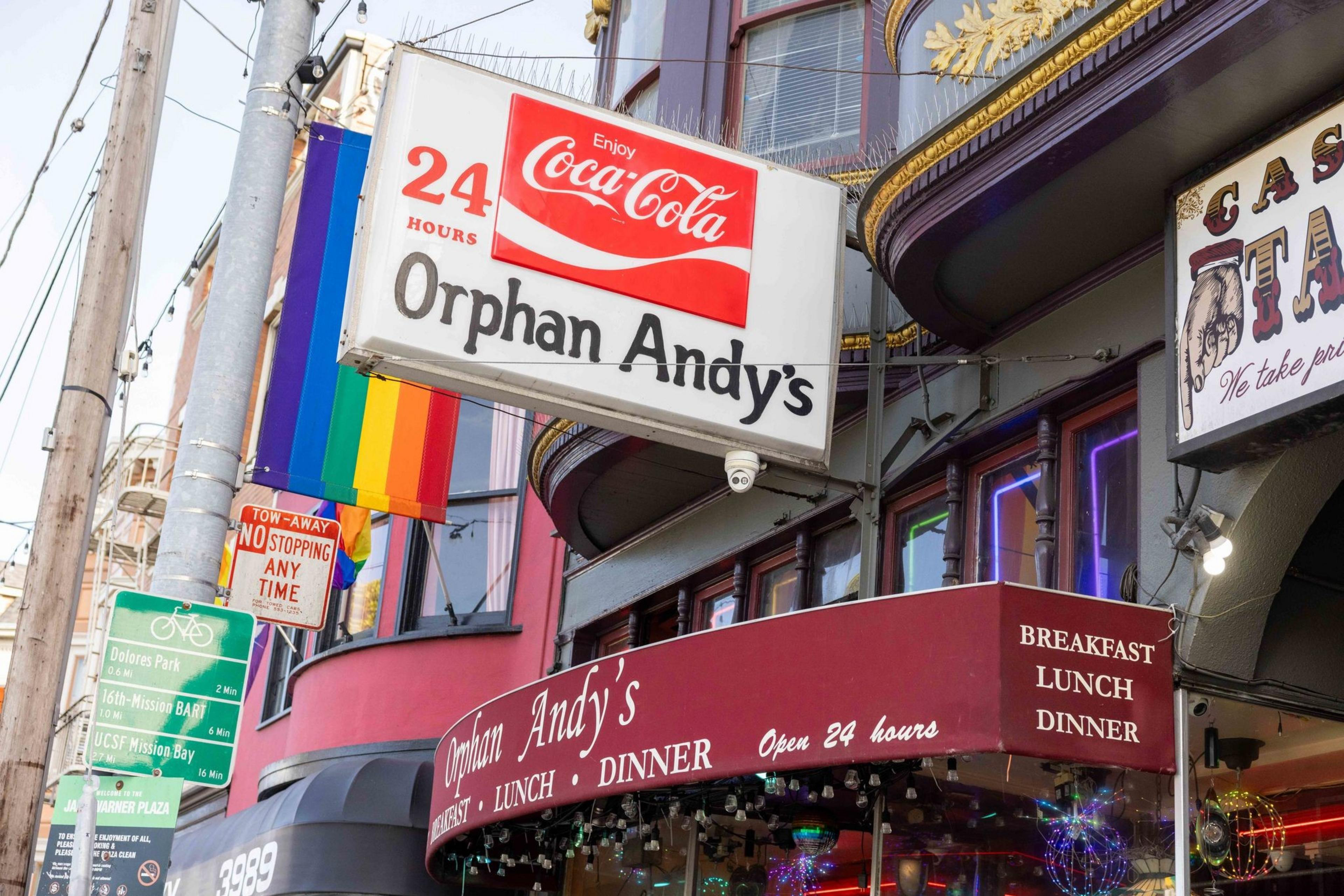 The image shows the entrance to "Orphan Andy's," a 24-hour diner. A rainbow flag is displayed prominently. Signs indicate it's open for breakfast, lunch, and dinner.