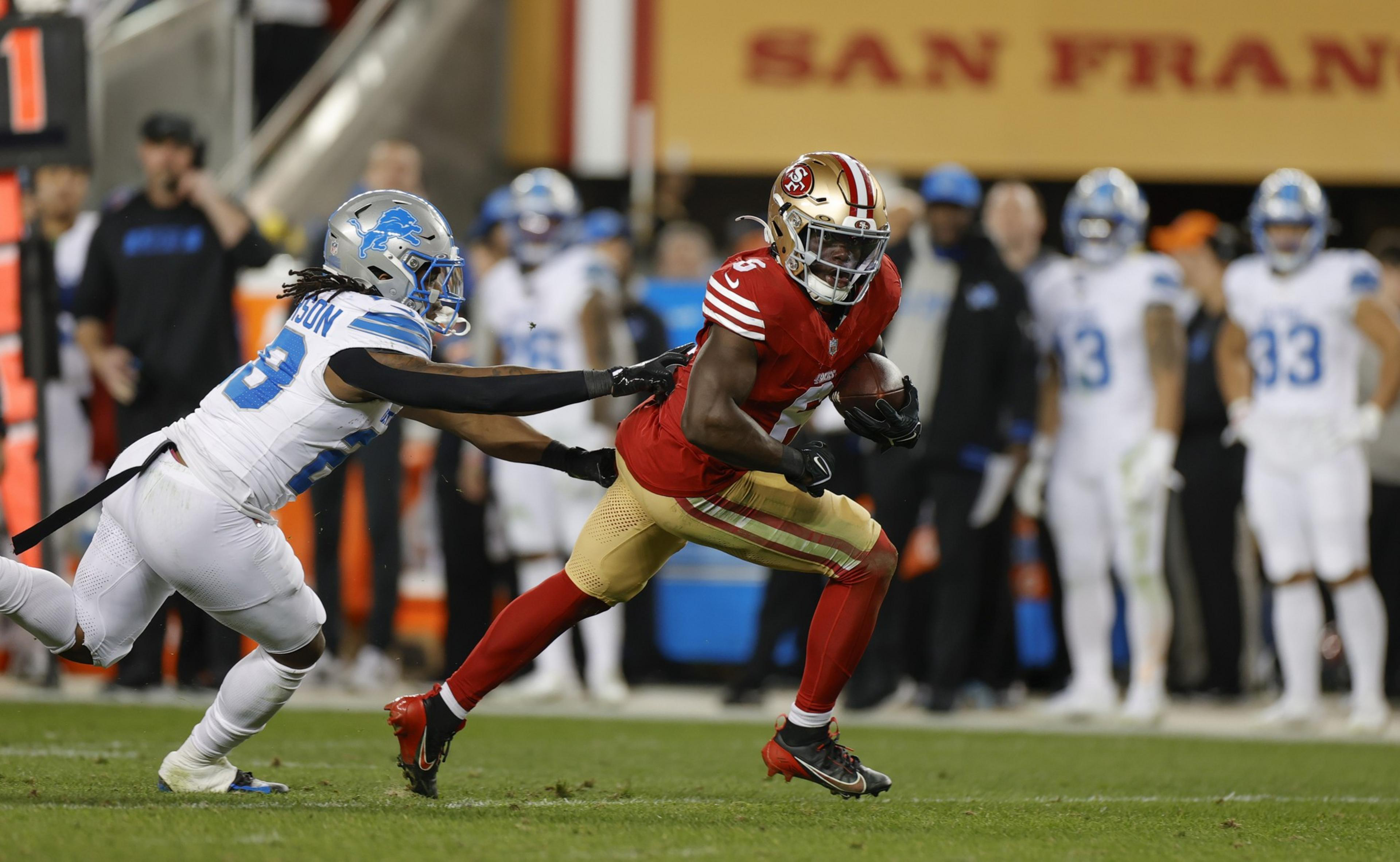 A San Francisco 49ers player runs with the ball while a Detroit Lions defender tries to grab his jersey during a football game.