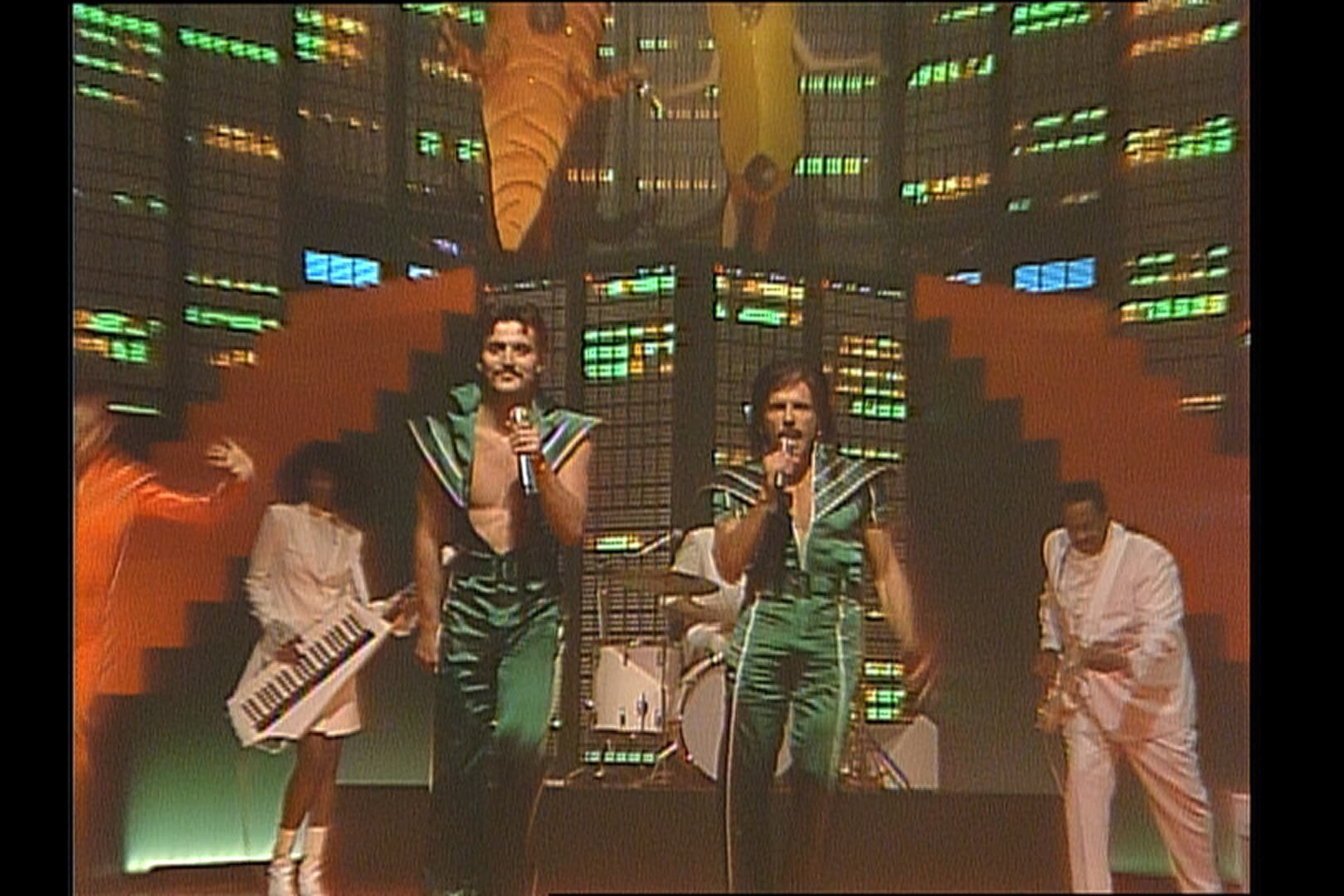 Two men in green shiny outfits sing into microphones while band members play keyboard and guitar against a colorful, futuristic backdrop.