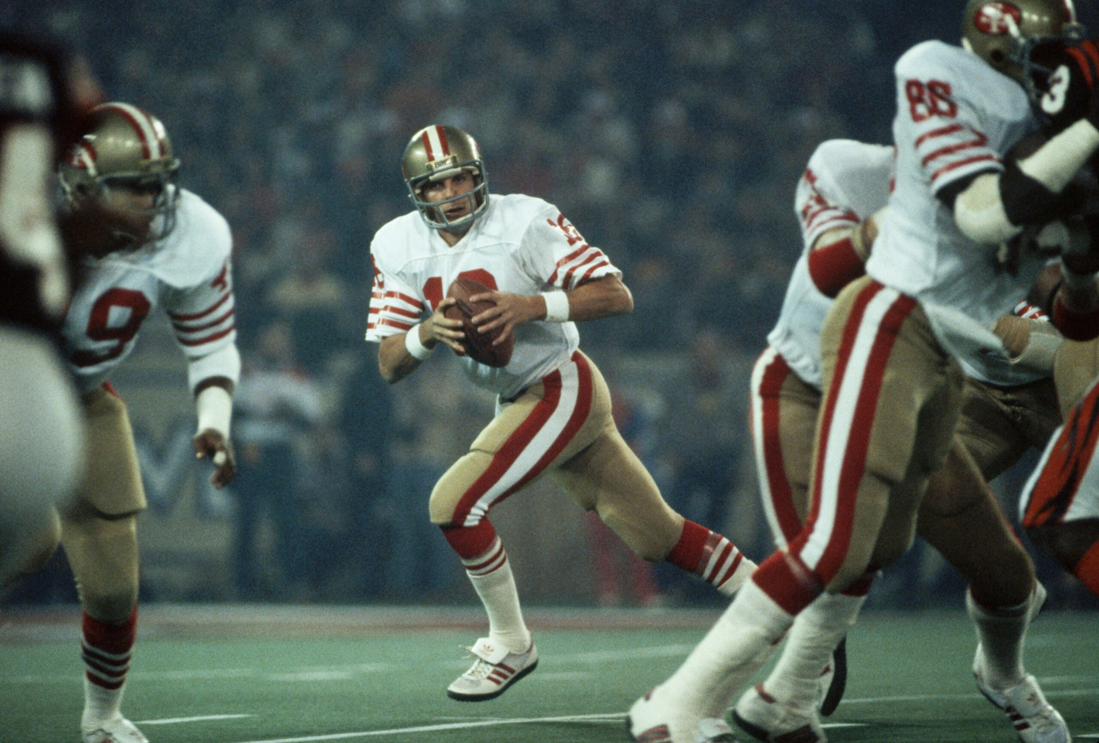 A football quarterback in a white and gold uniform prepares to throw the ball while teammates block opponents on the field.