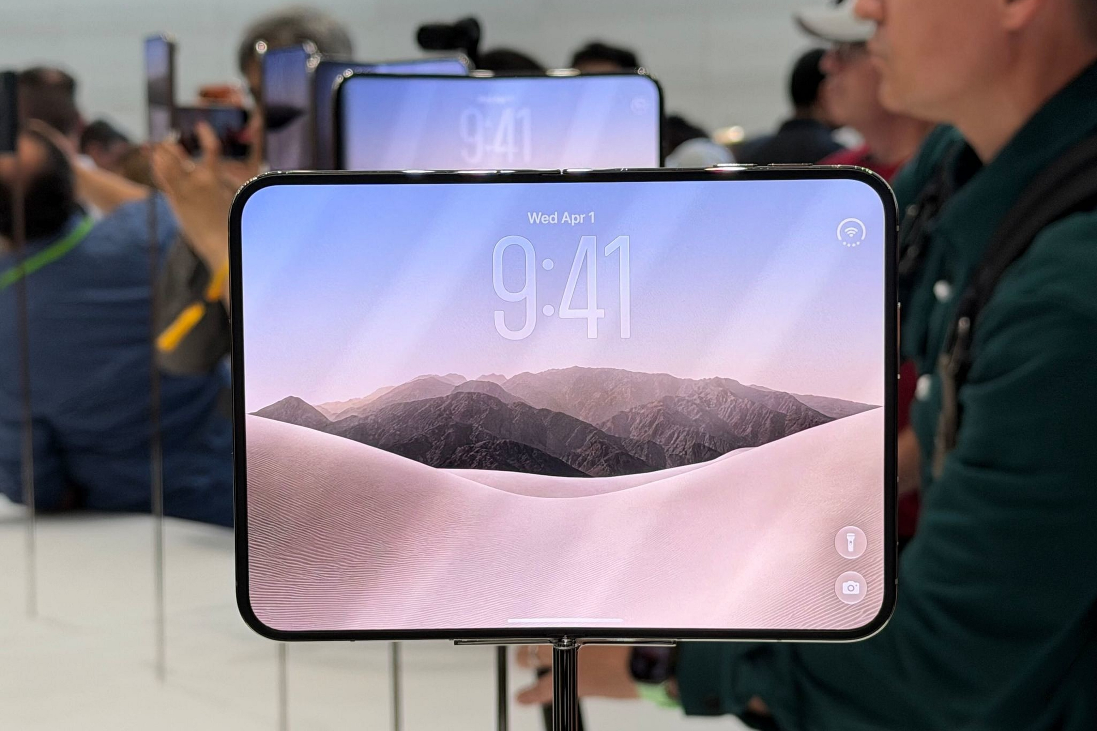 A horizontally oriented smartphone with a landscape wallpaper displays the time 9:41 and date Wed Apr 1, mounted on a stand with people and reflections behind it.