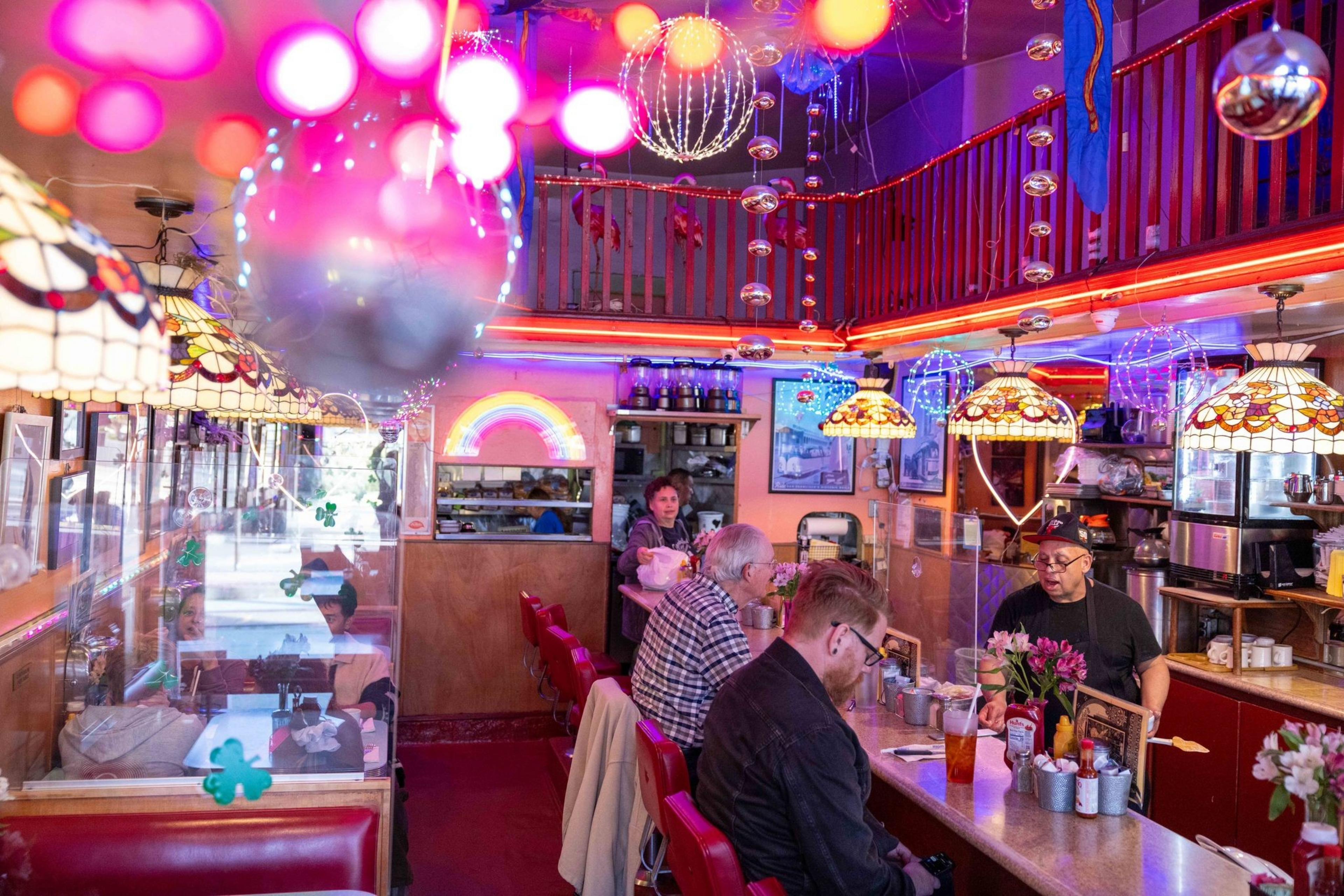The image shows a colorful diner with red seating, vibrant hanging lamps, and people dining. A staff member is behind the counter. Neon lights and decorations adorn the space.