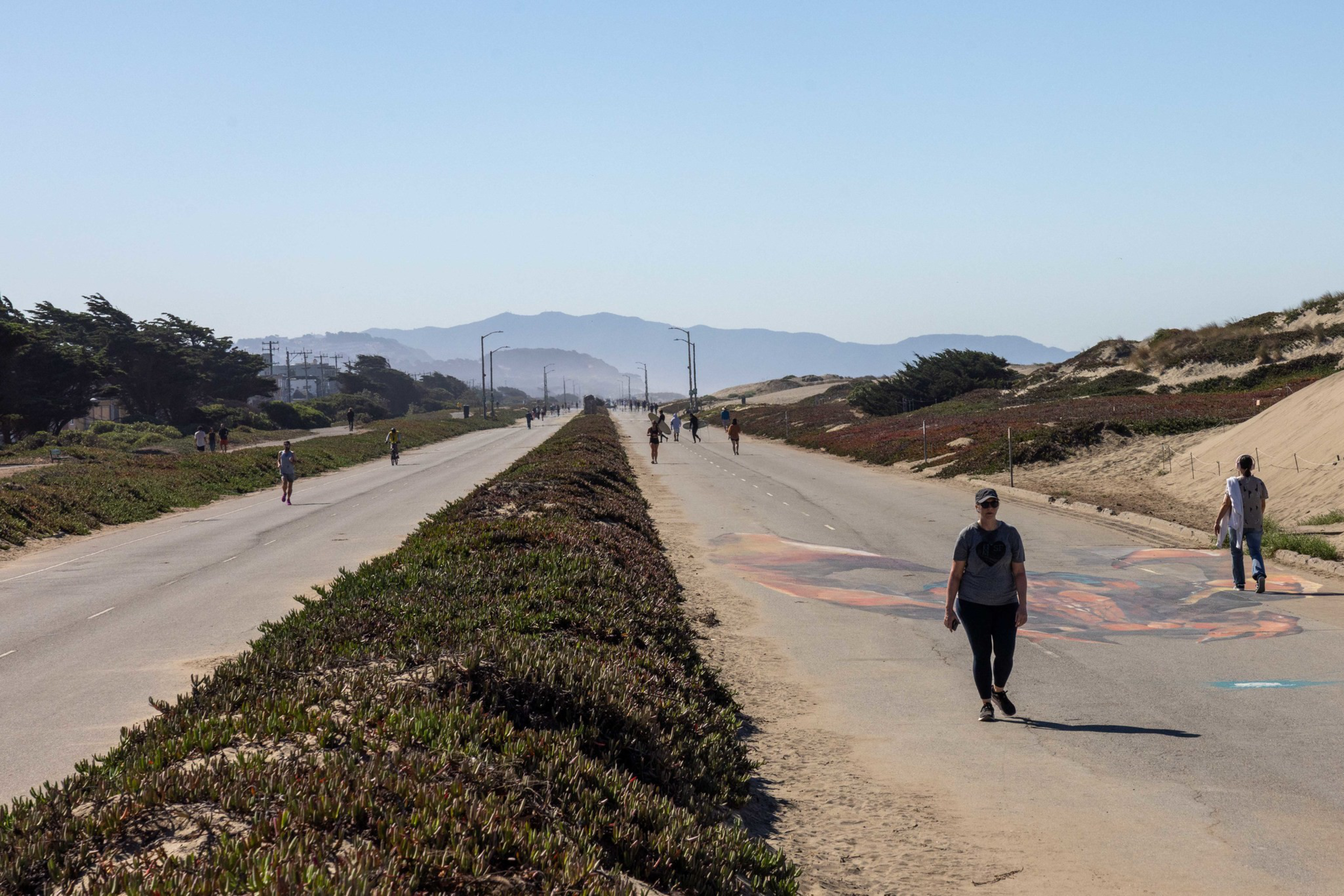 A wide, empty road divided by greenery shows people walking, biking, and jogging with dunes and mountains in the distant background under a clear sky.