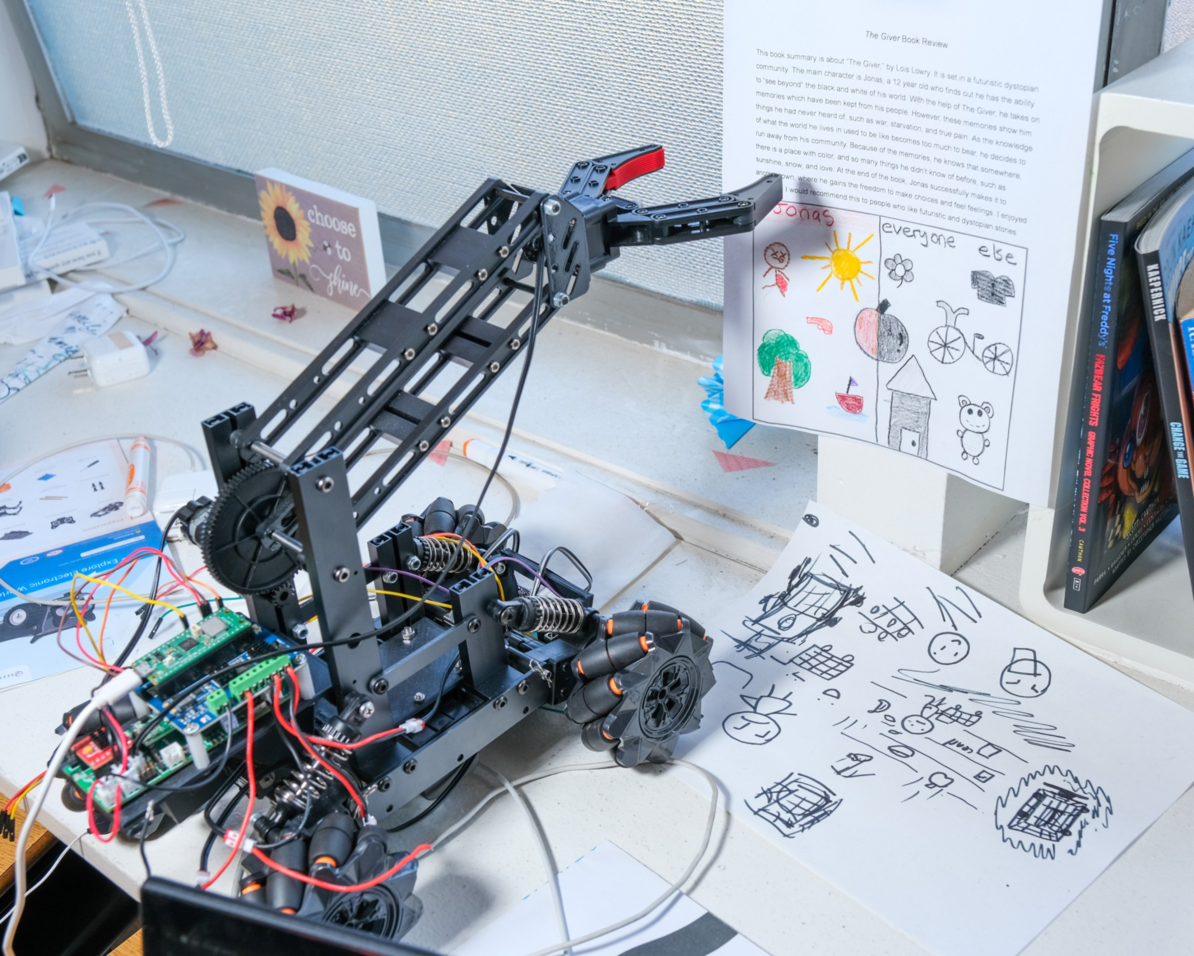 A black mechanical robot arm with exposed circuitry and colorful wires grabs a child's drawing from a cluttered desk with papers and books in the background.