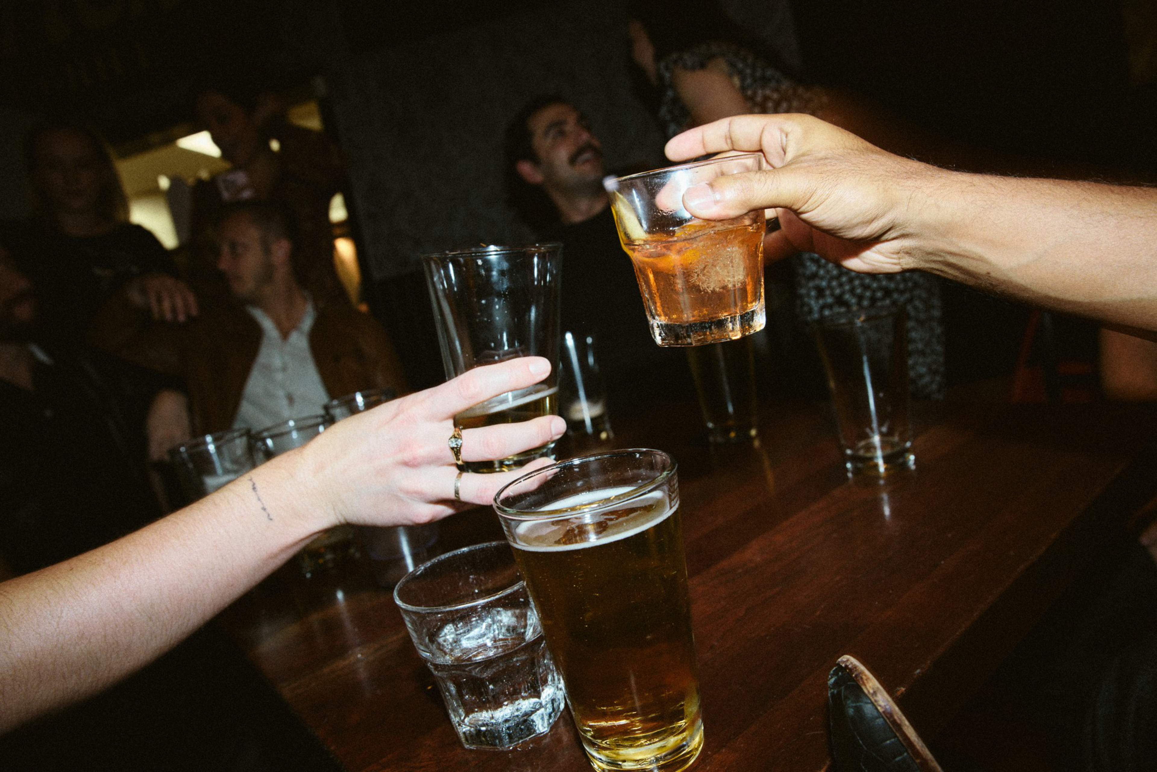 Two hands clink glasses over a wooden table with various drinks, while a group of people socializes in the dimly lit background.