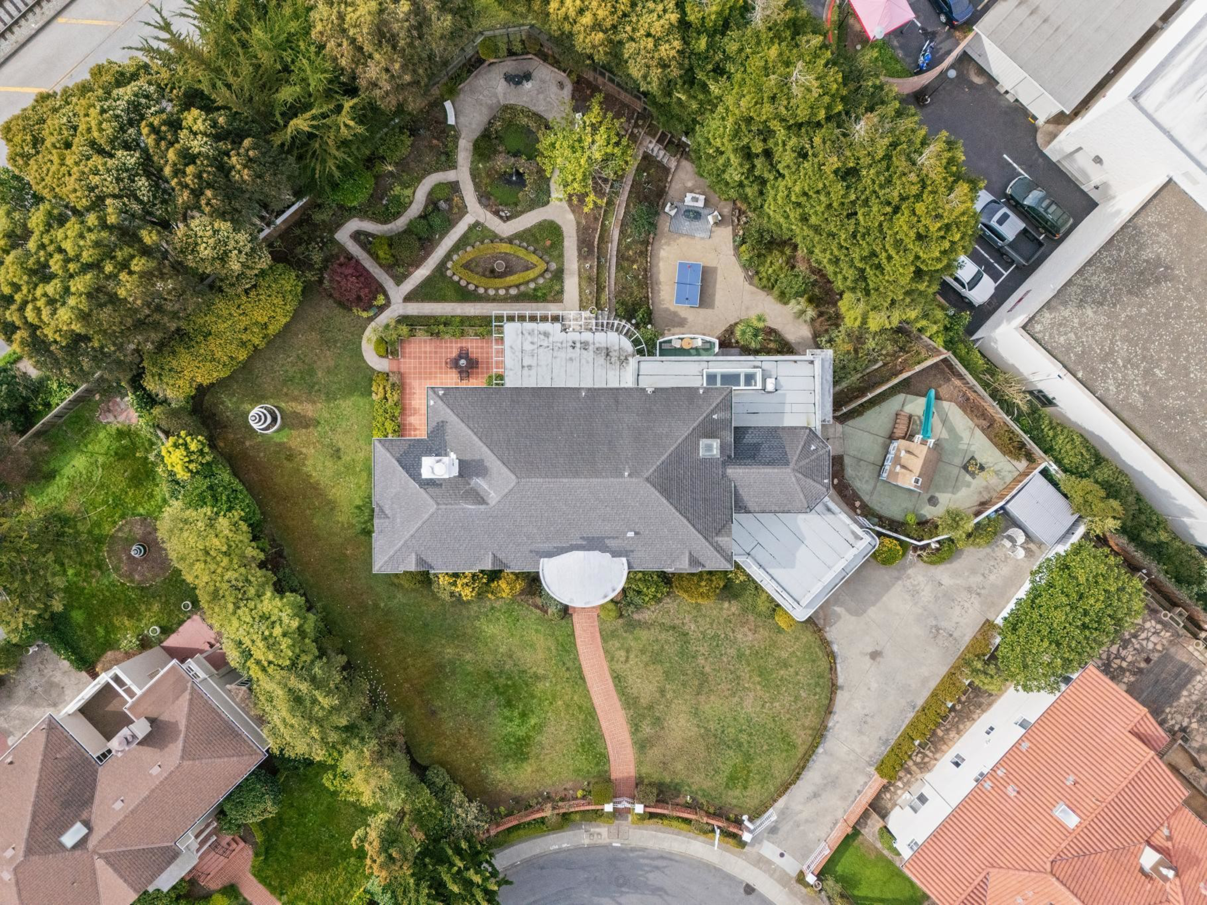 A large house with a gray roof sits in the center of a green yard, featuring curved walkways, a garden, a playground, and surrounding trees and neighboring buildings.