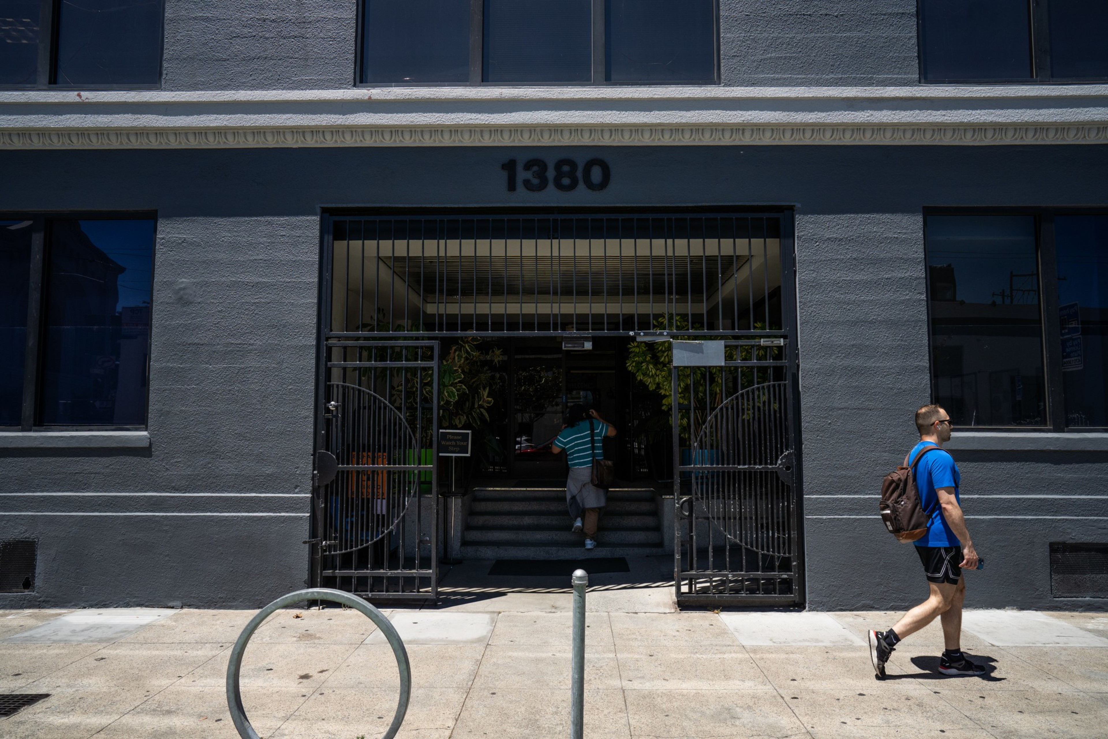 A gray building with the number 1380 above a gated entrance. One person walks on the sidewalk, while another person enters the building.