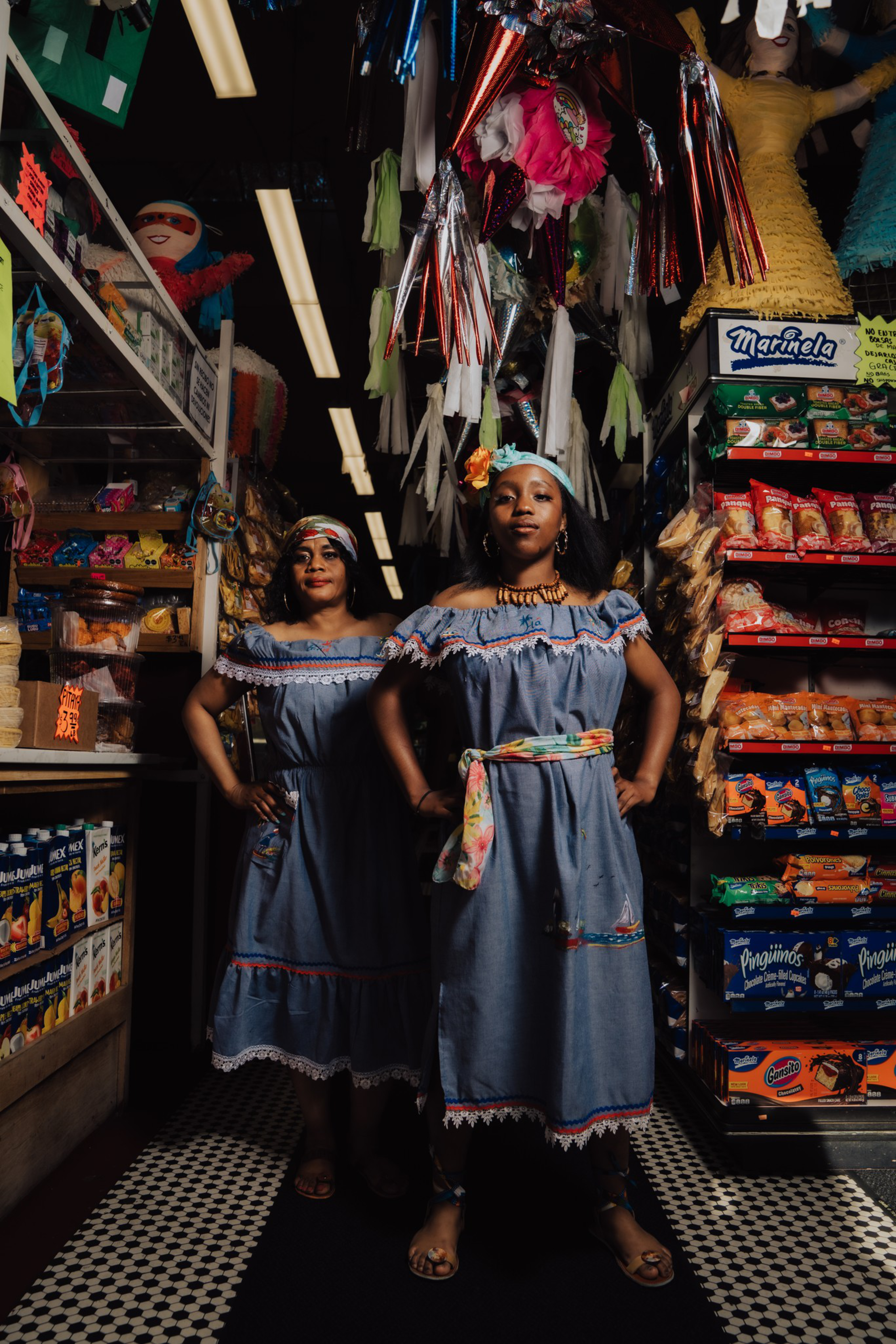 Two women in matching blue off-shoulder dresses with floral accessories stand confidently inside a colorful store filled with snacks and piñatas.