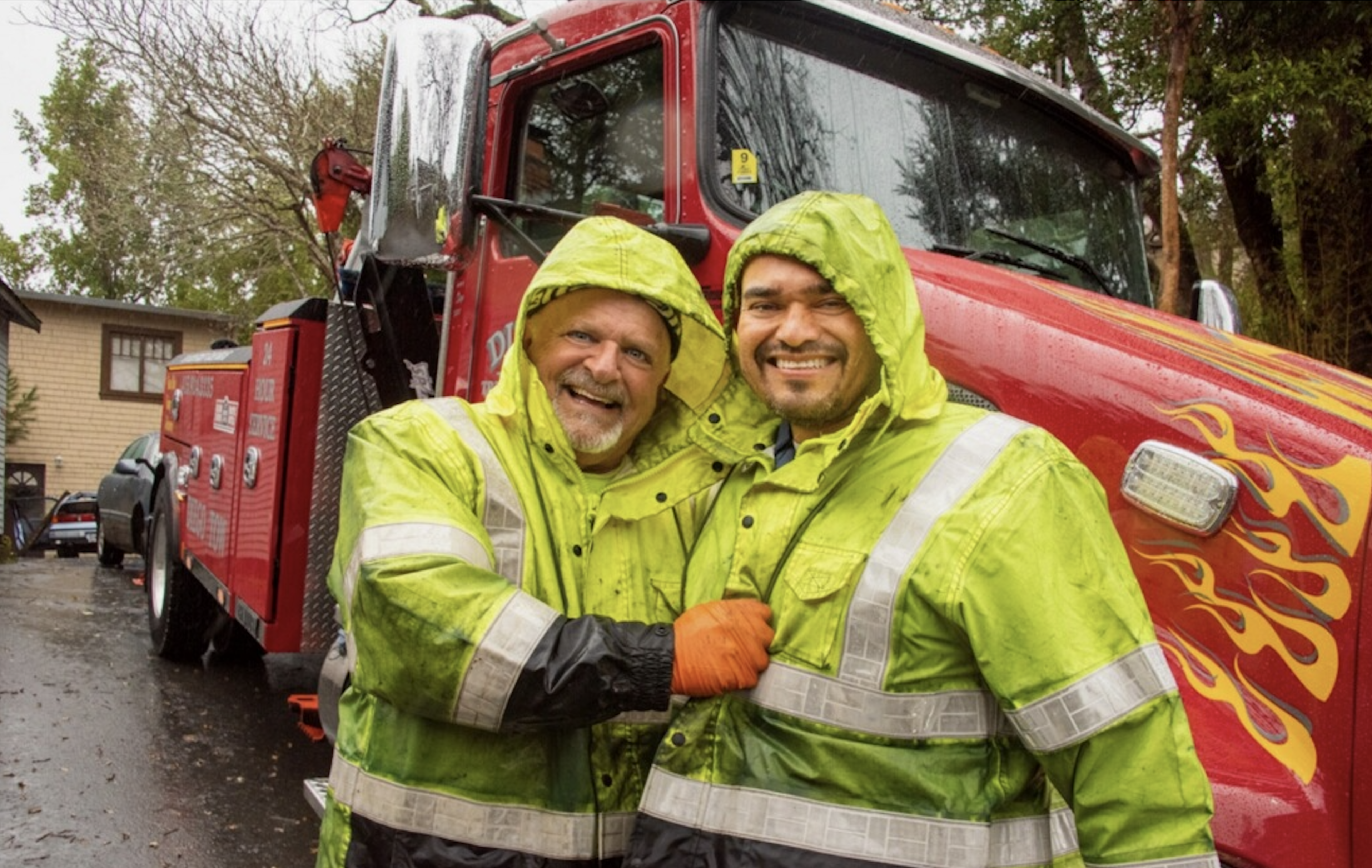 Two men wearing bright yellow rain jackets smile and pose in front of a red tow truck with flame decals on a rainy day.