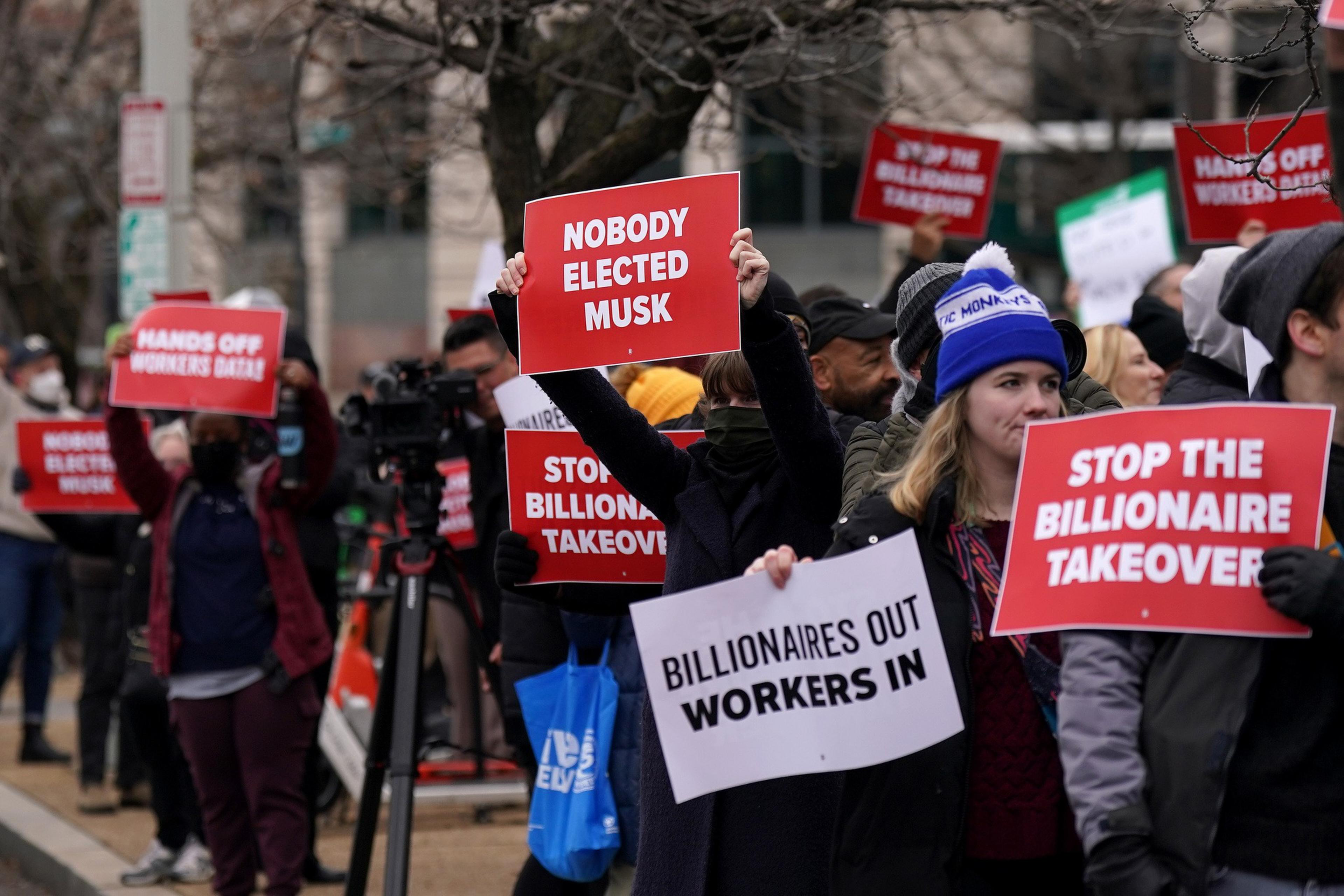 People are gathered, holding signs with messages like "NOBODY ELECTED MUSK" and "STOP THE BILLIONAIRE TAKEOVER," protesting against corporate influence.