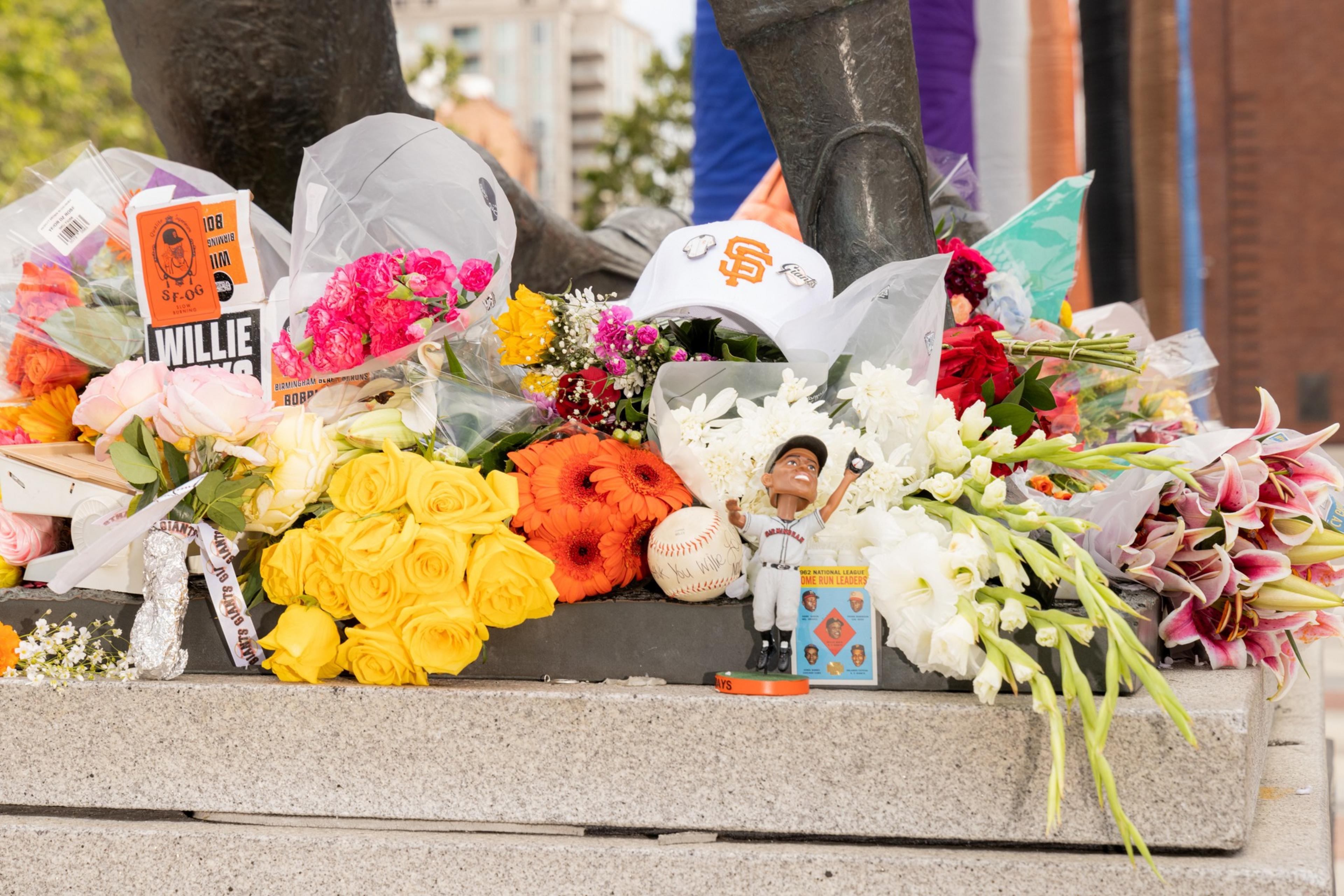 The image shows a memorial with various flower bouquets, a white baseball cap, a baseball, and a figurine of a baseball player. The flowers are vibrant and colorful.
