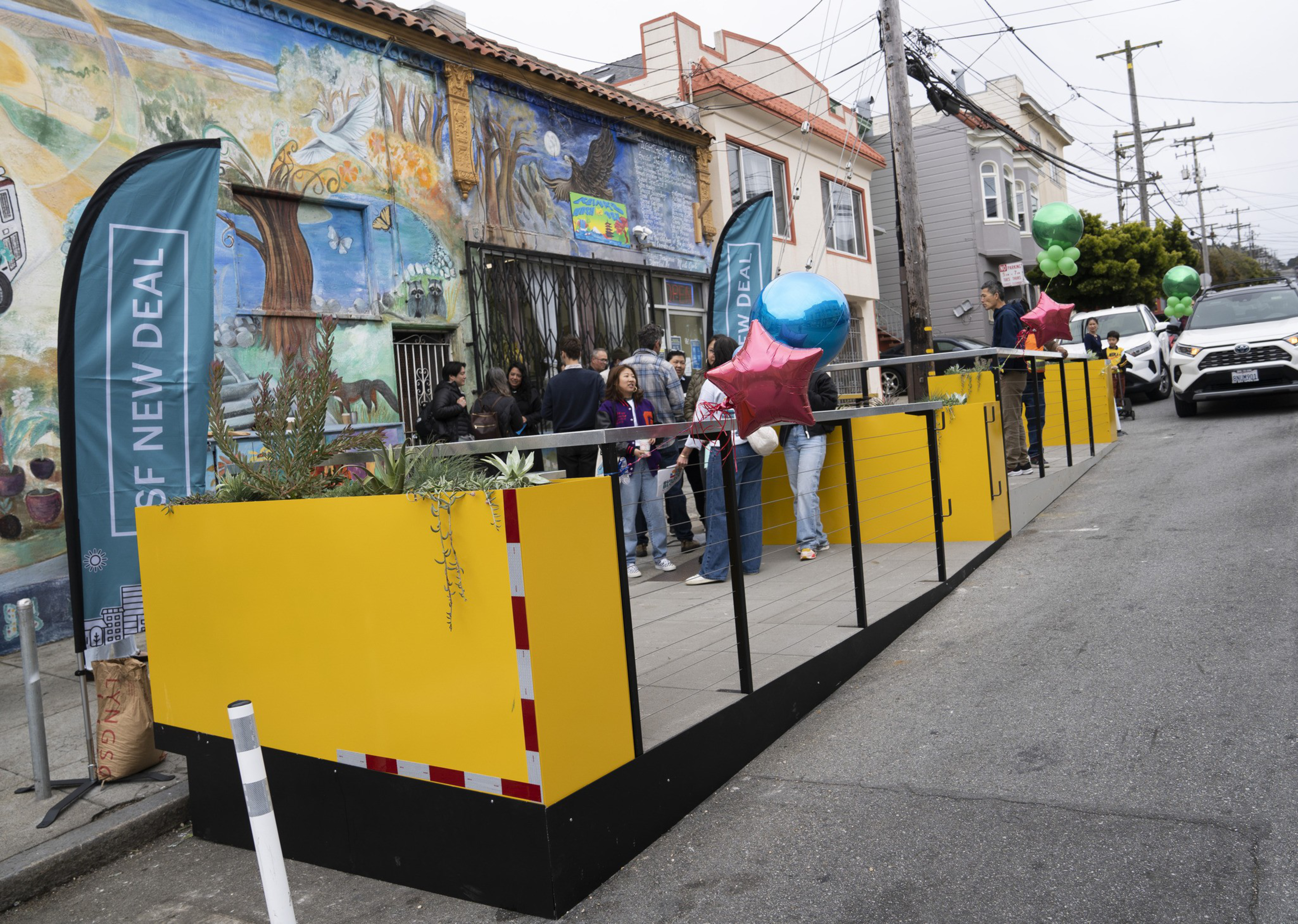 A brightly painted mural serves as the backdrop for a group of people gathered on a sidewalk parklet. Yellow barriers, plants, and colorful balloons decorate the area.