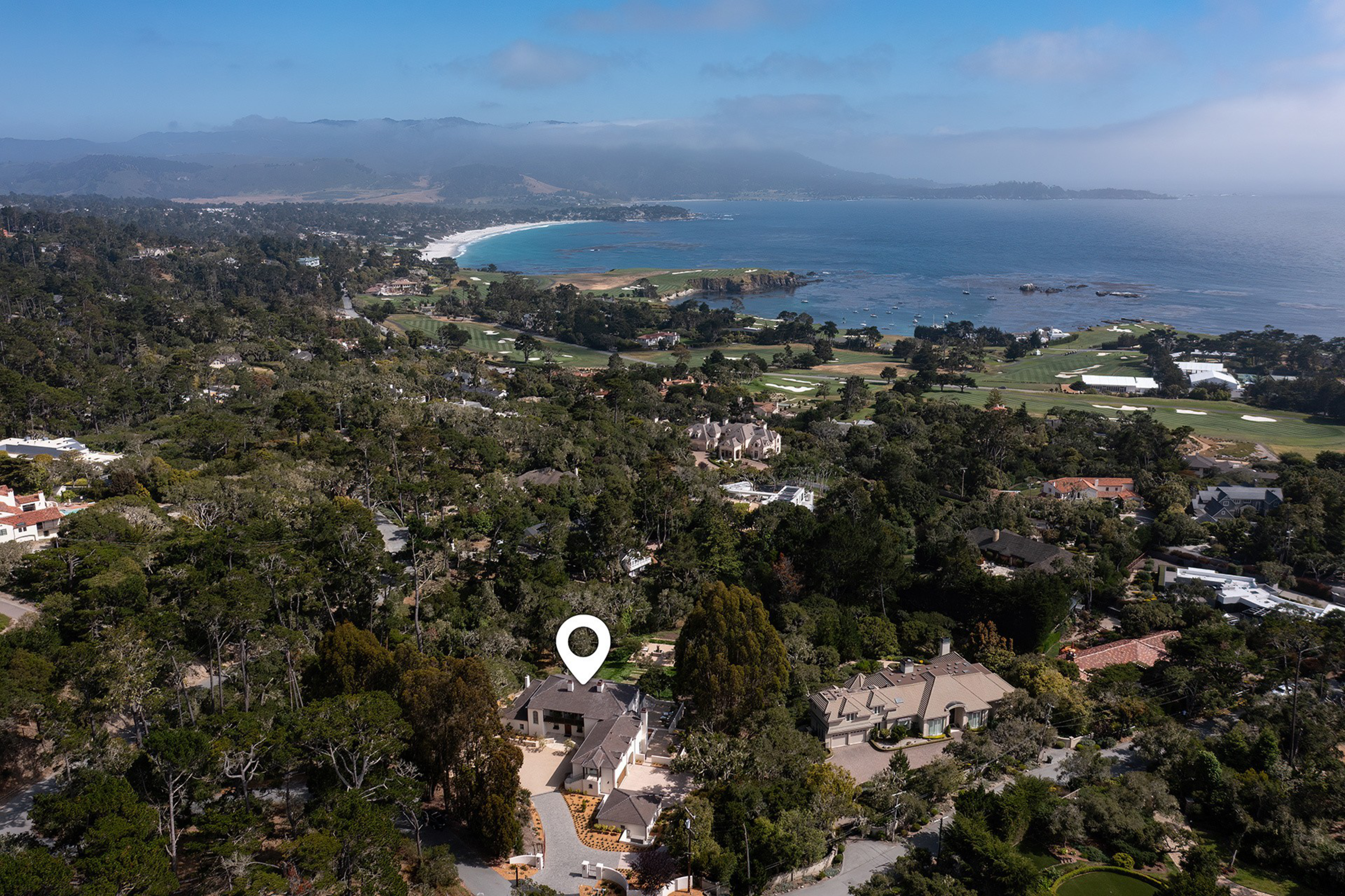 A coastal town with dense trees, several houses, a golf course, and a bay with sandy beaches and mist-covered hills in the distance.