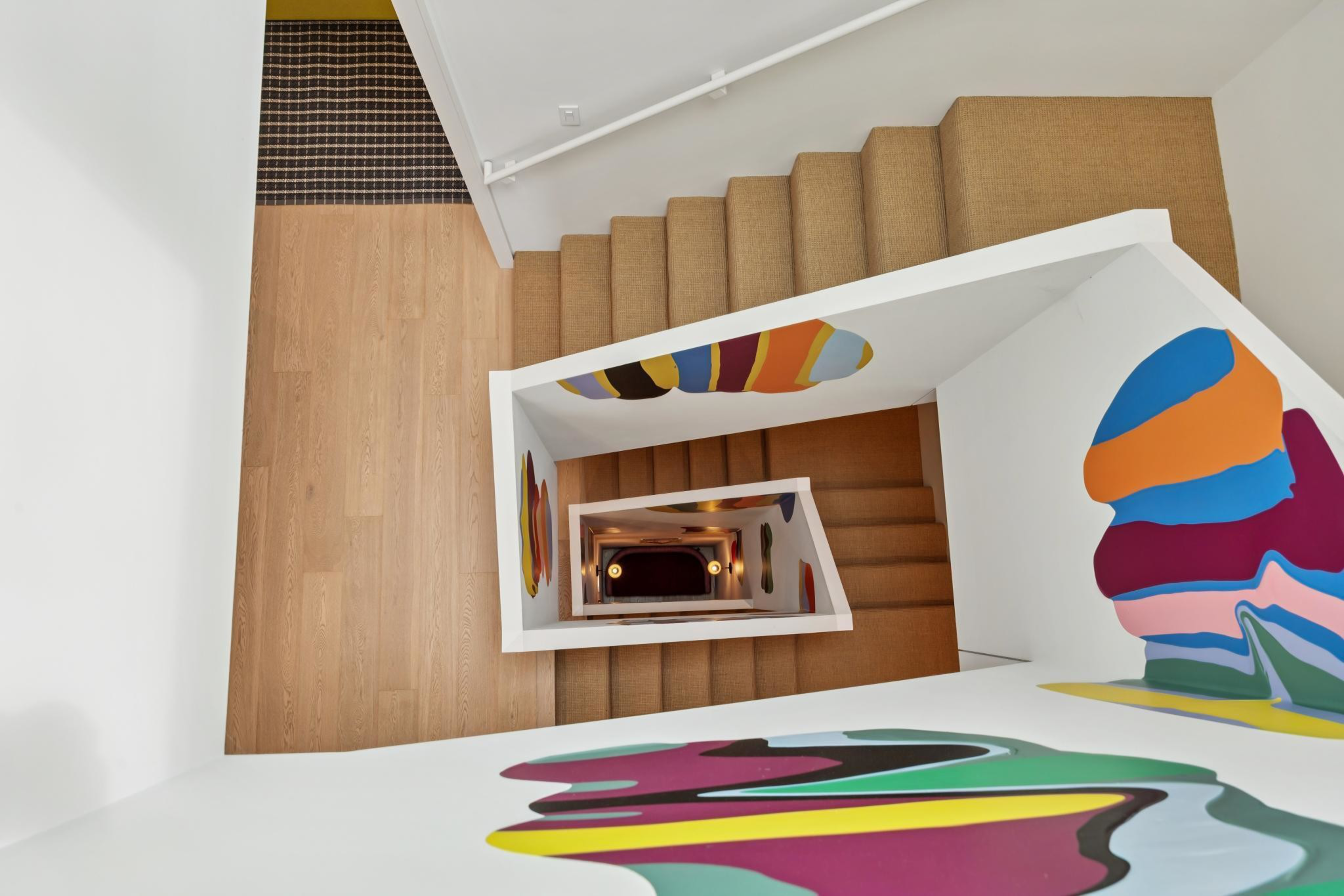A spiral staircase with natural wood steps and white walls features colorful abstract designs on the walls, viewed from the top looking down.