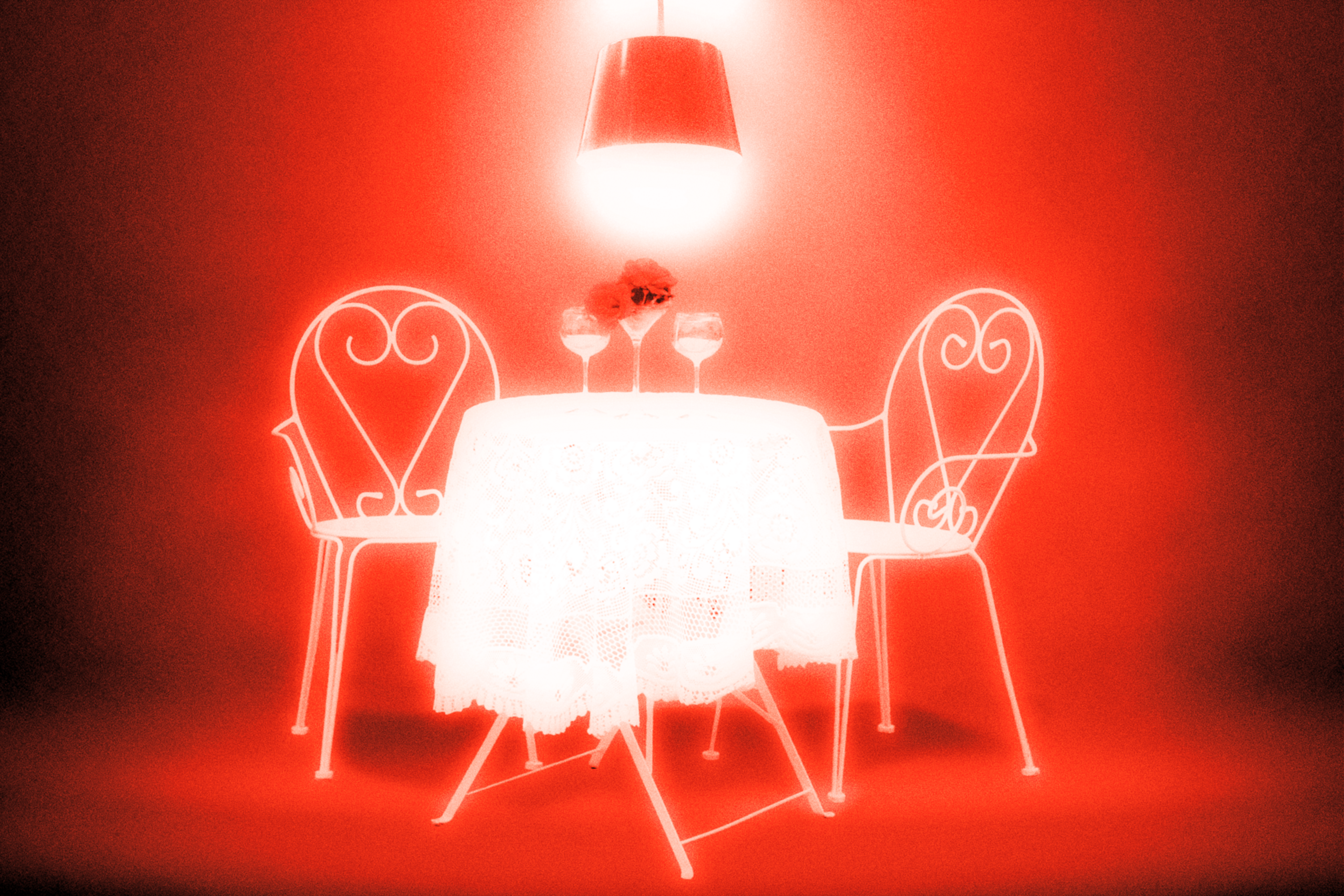 A small, round table with a lace tablecloth, two empty wine glasses, and a vase with red flowers sits between two ornate metal chairs under a hanging lamp.