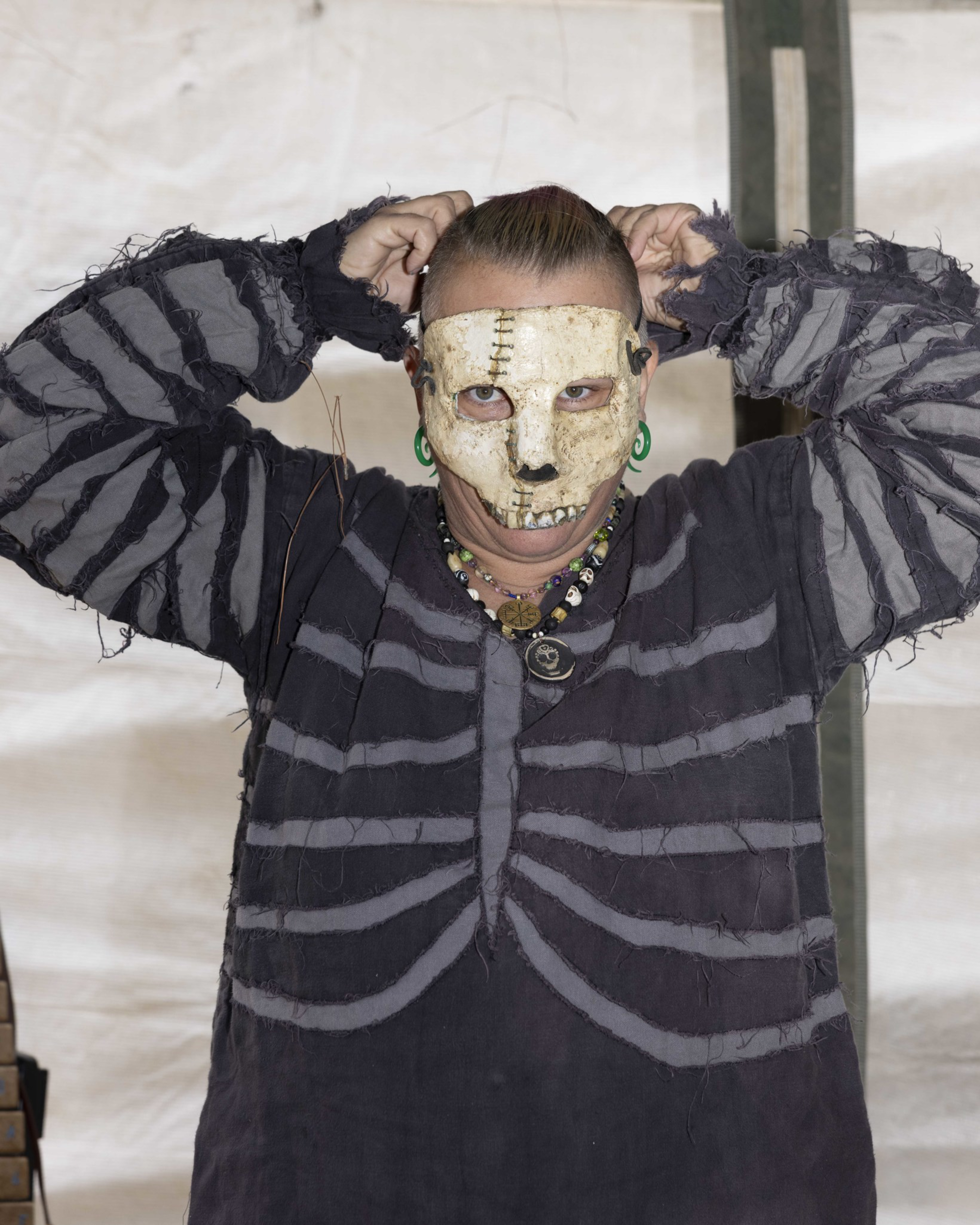 The individual is wearing a skeleton-themed costume with a stitched white skull mask and adjusting their hair. They also have green ear plugs and multiple necklaces.