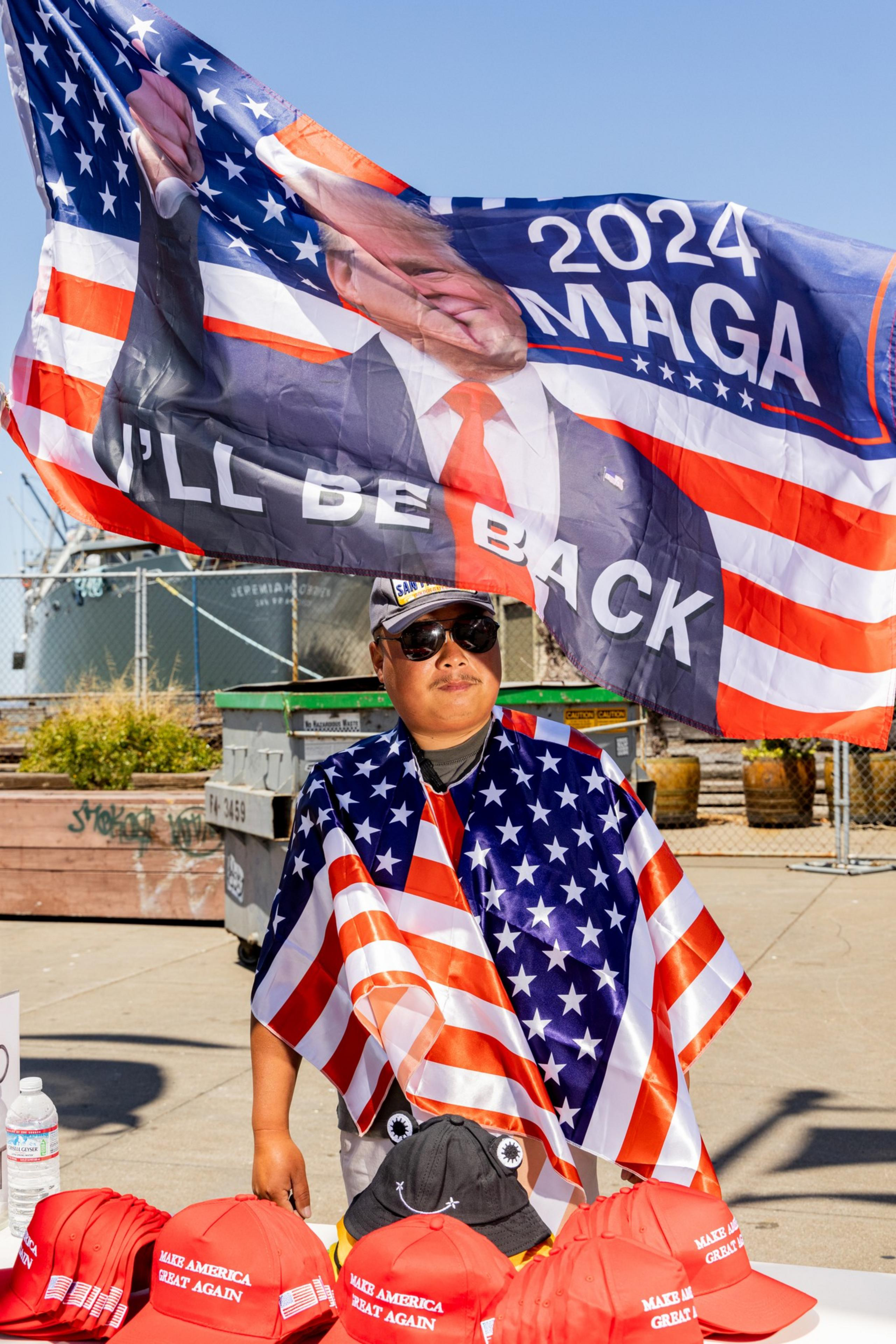 A person wearing sunglasses and draped in an American flag stands in front of a large 2024 "MAGA" flag, with several red "Make America Great Again" hats on display.