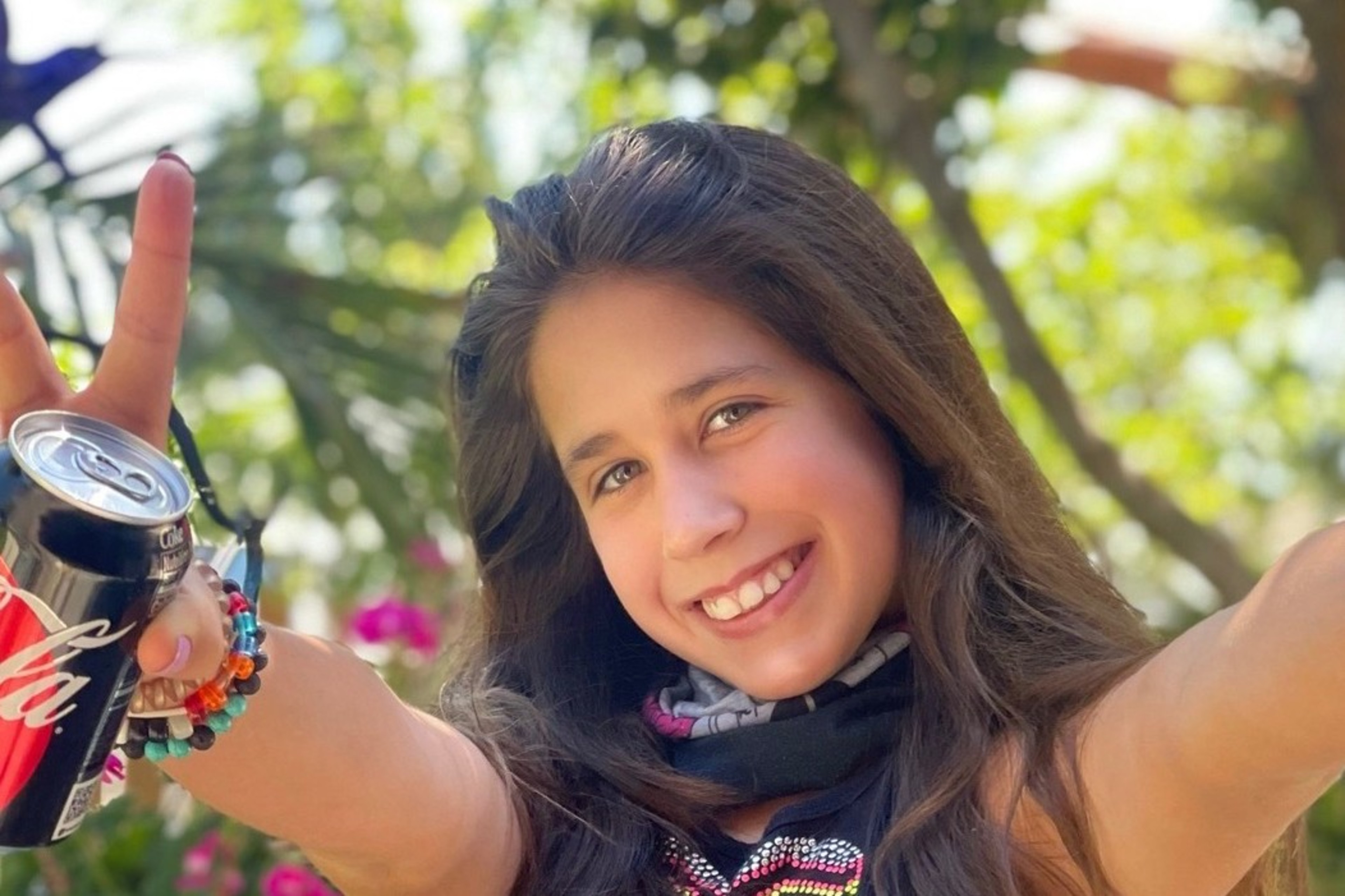 A smiling girl holds a Coke can, making a "peace" sign, with blurry greenery behind her.
