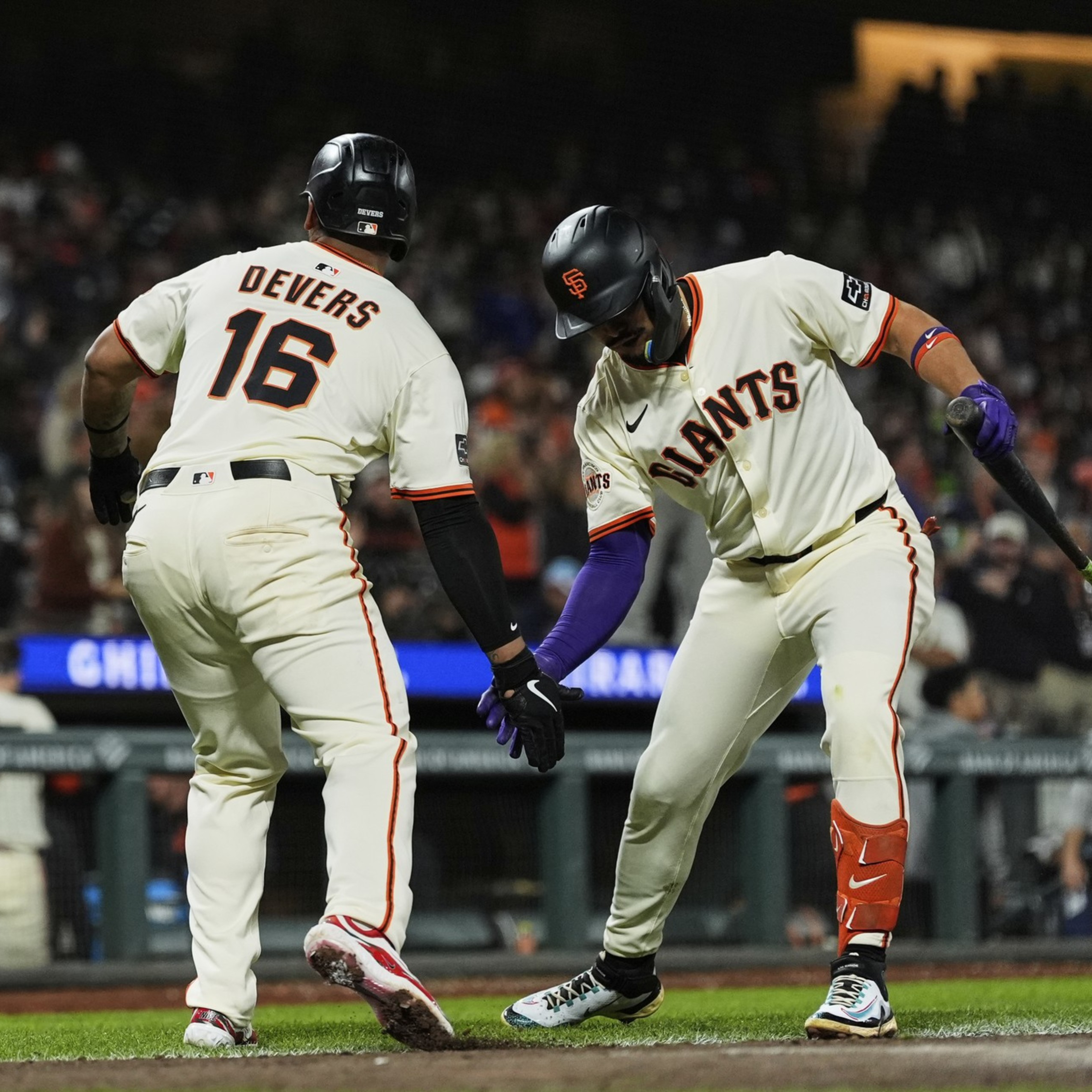 Two baseball players from the Giants, one named Devers, are giving a low handshake on the field during a game with fans in the background.