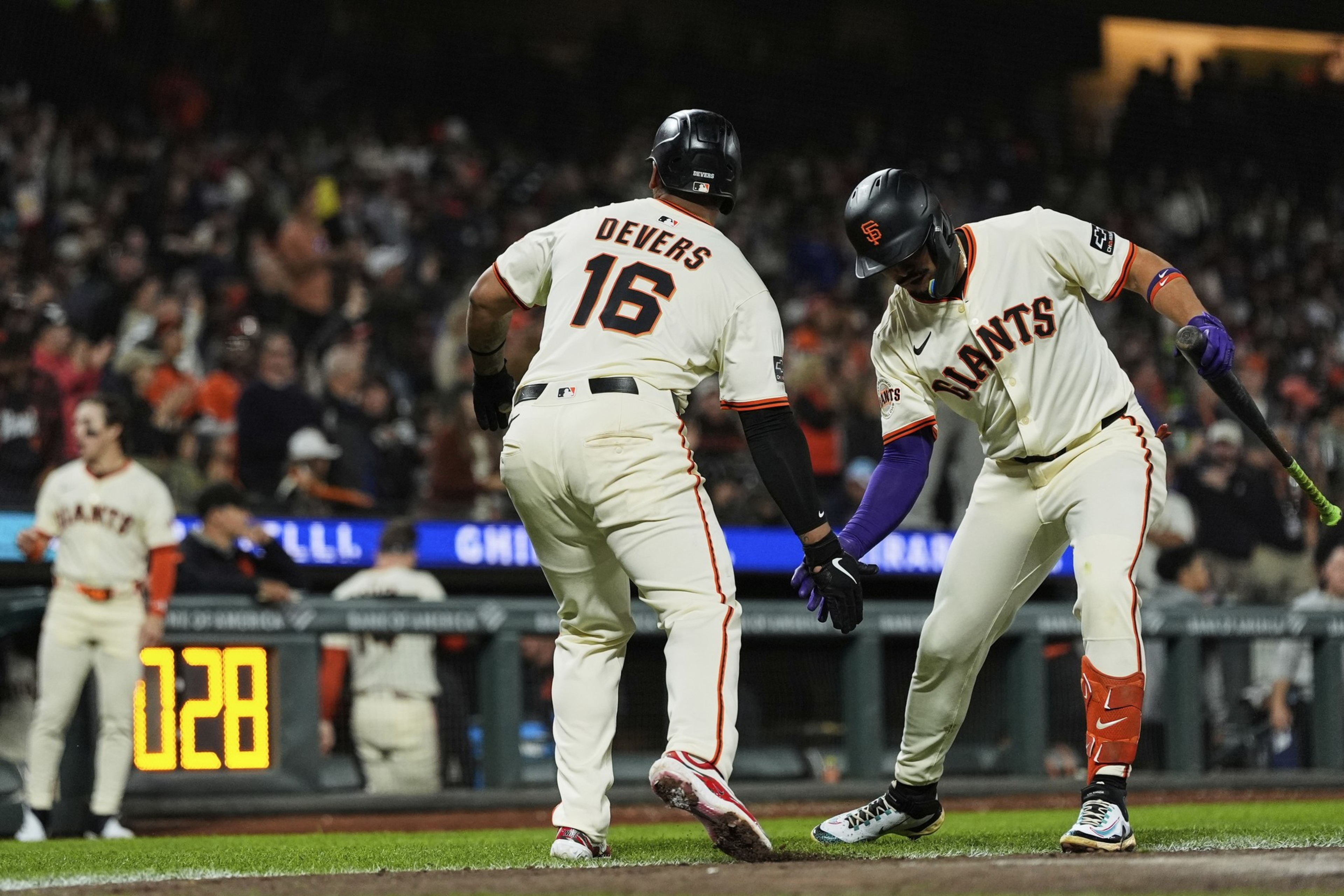 Two baseball players from the Giants, one named Devers, are giving a low handshake on the field during a game with fans in the background.