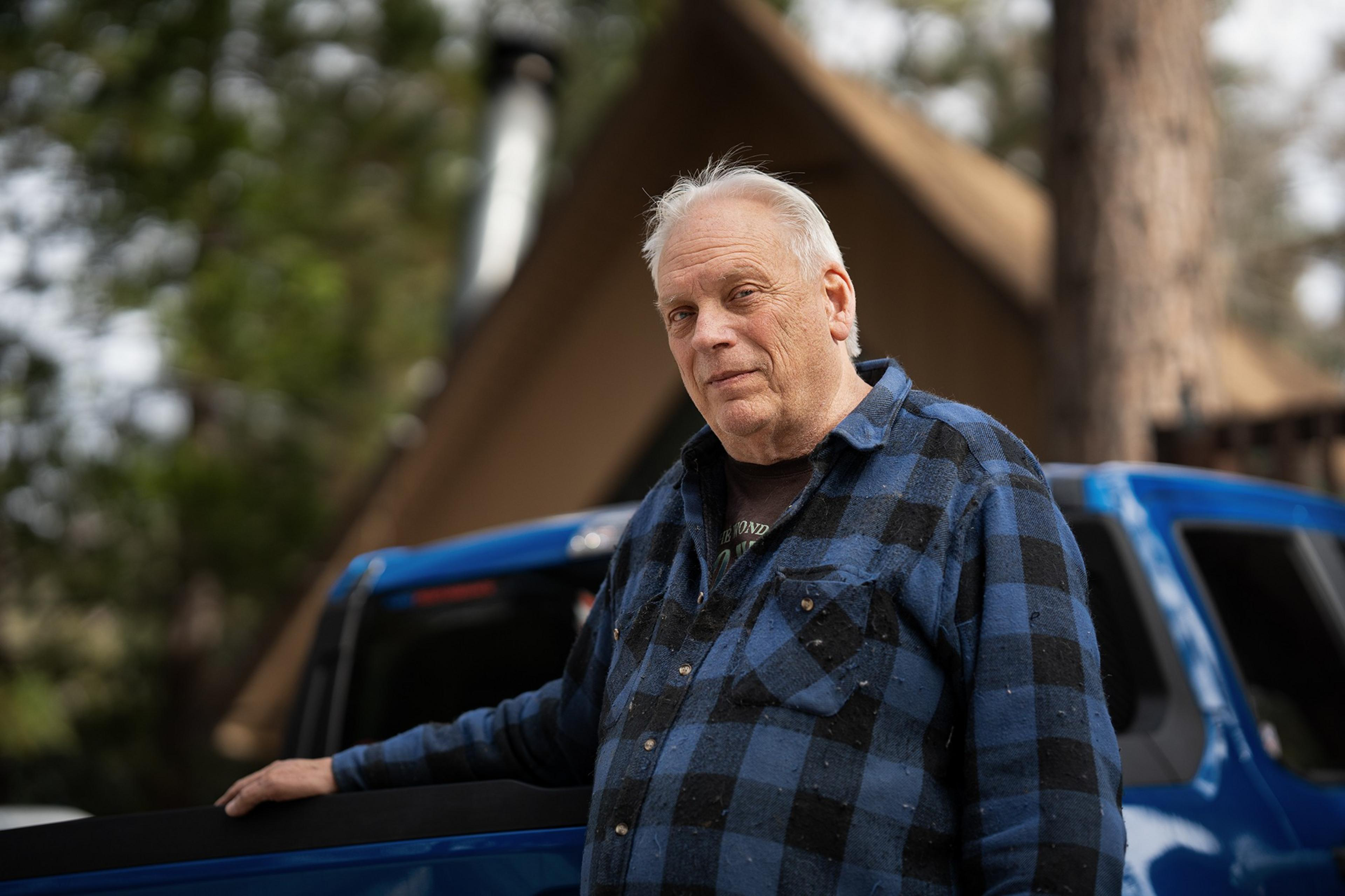 Man in blue and black plaid fanel stnds in front of blue pick up truck and a frame home