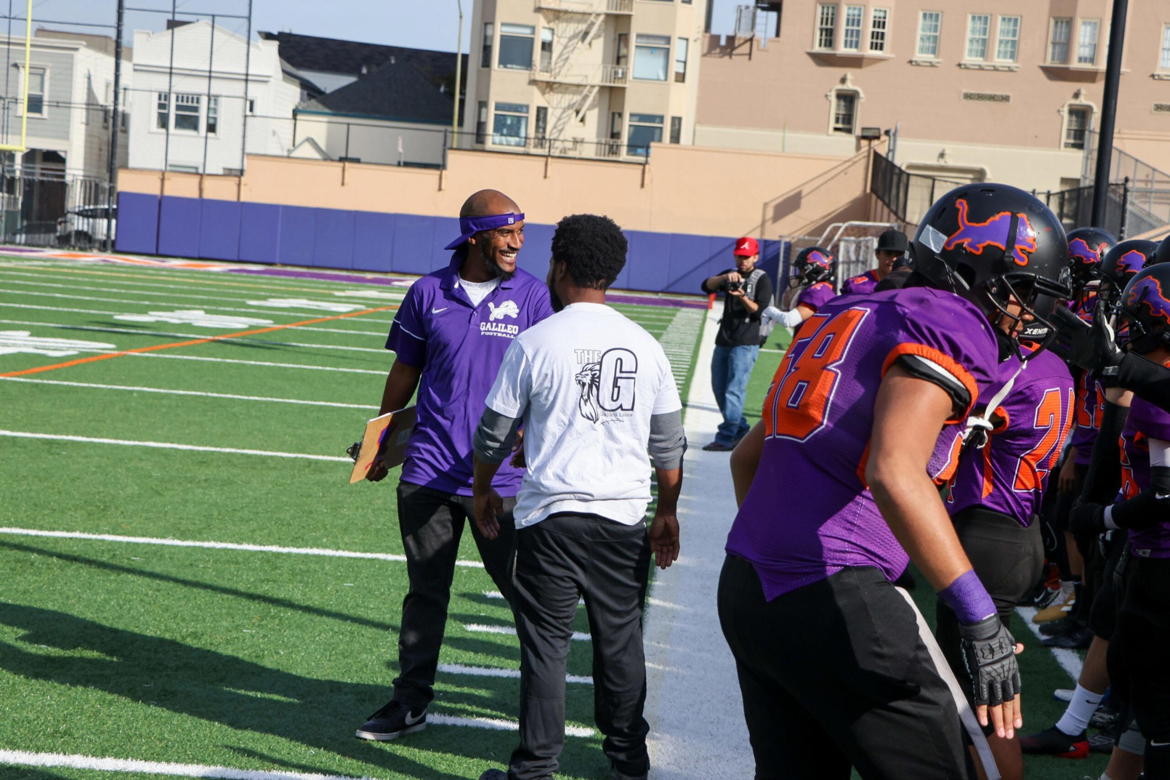 A football coach in purple talks with a man in a white shirt on the sidelines, while players in purple and orange uniforms prepare near the field.