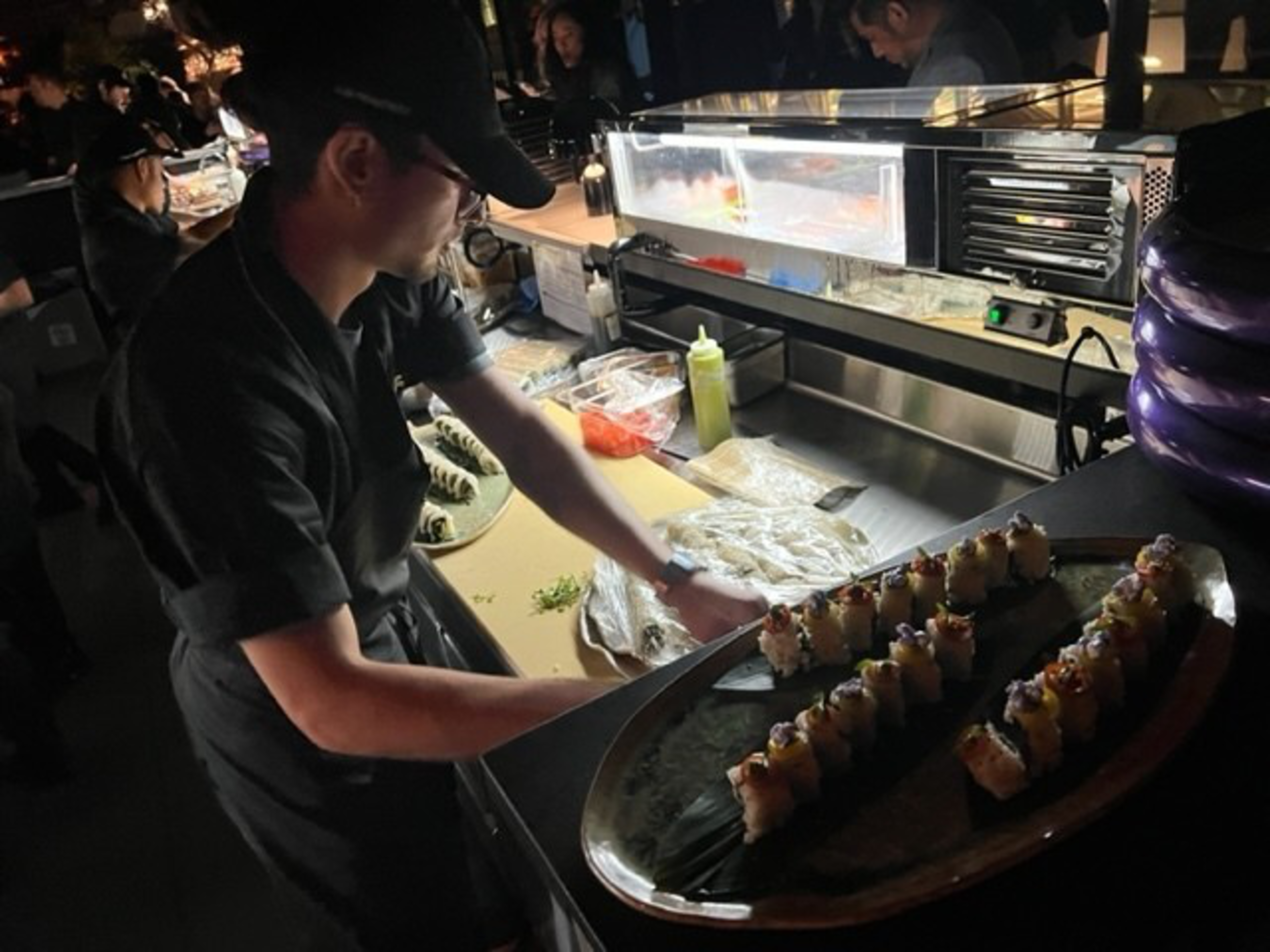 A chef in black attire arranges sushi rolls on a large platter in a dimly lit kitchen with other chefs working in the background.